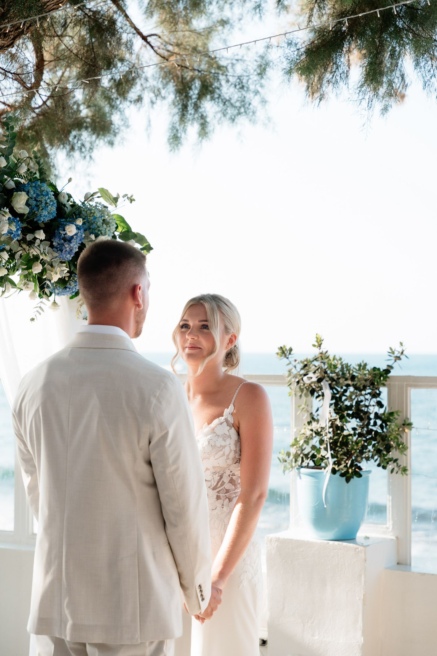 A couple stands together under floral decorations at a seaside wedding venue with white decor and ocean views.