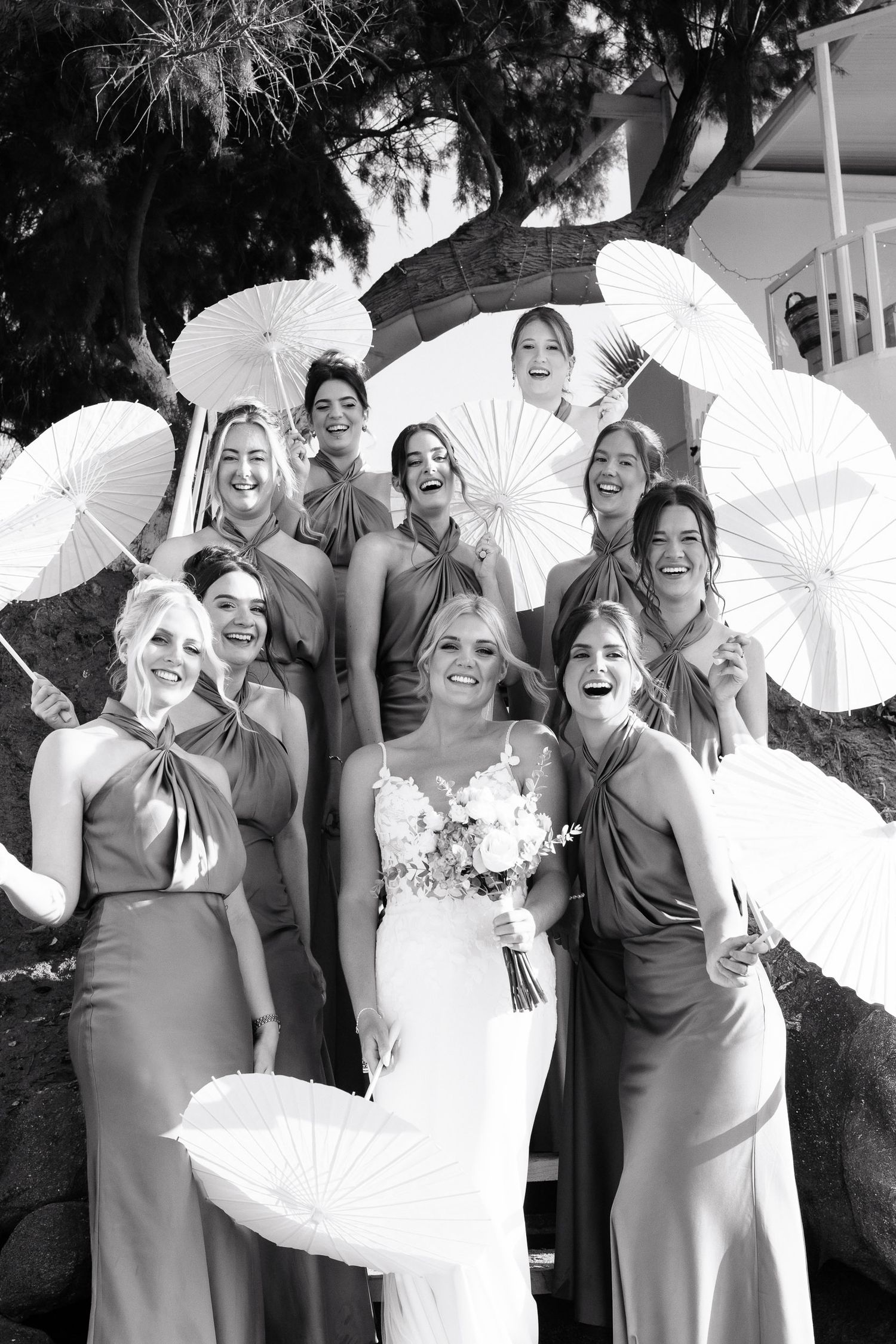 A wedding party poses with white parasols in a black and white photograph outdoors.