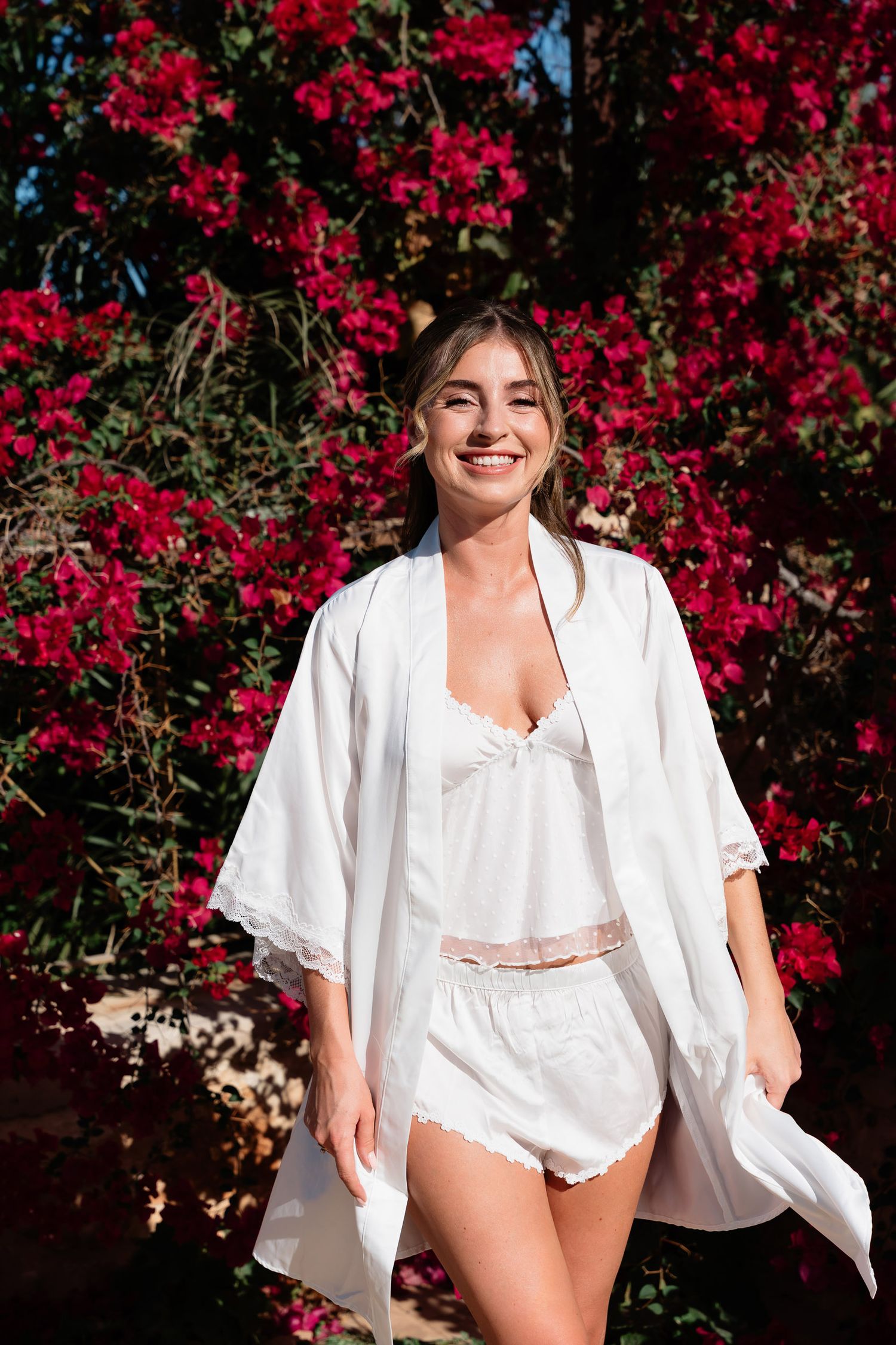 A person in a flowing white outfit poses against a backdrop of vibrant red bougainvillea flowers in a garden setting.