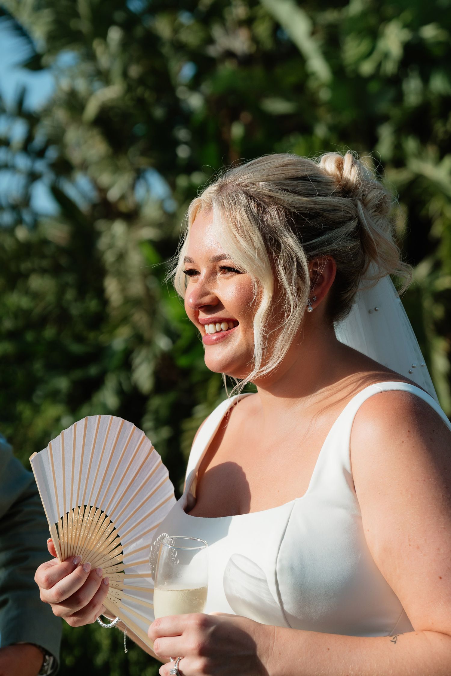 A bride in a white dress smiles while holding a folding fan and drink glass on a sunny outdoor day.