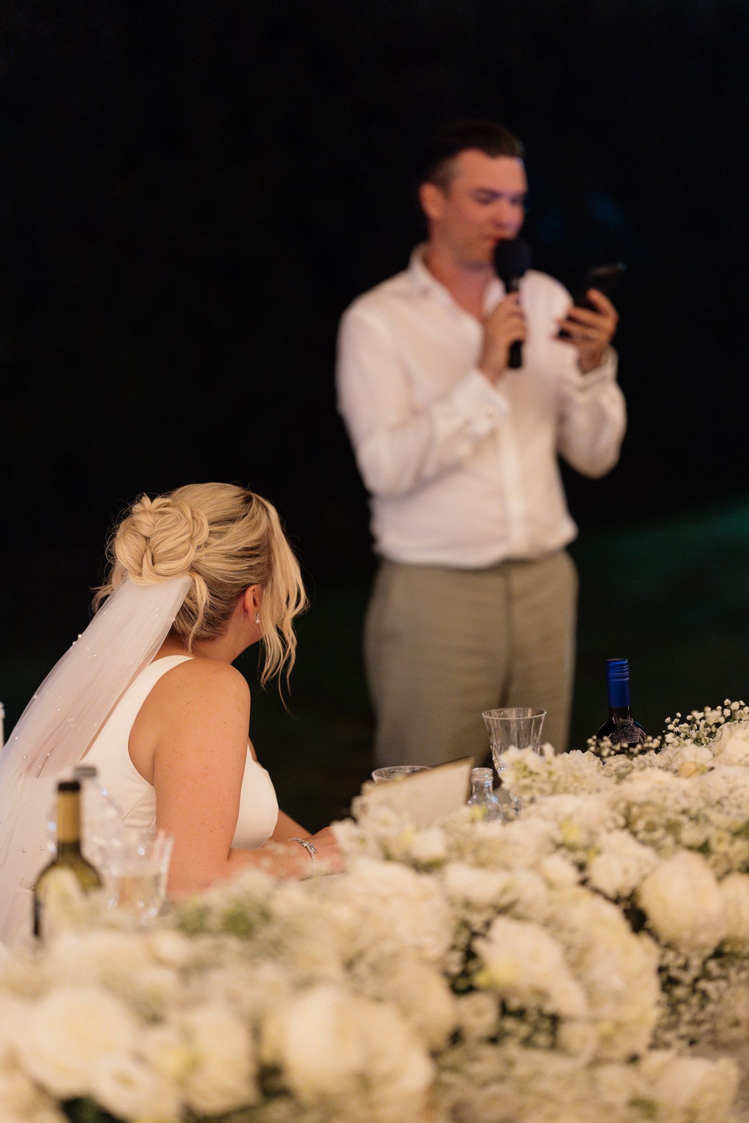 A wedding reception table decorated with white roses and baby's breath flowers while guests sit in the background.