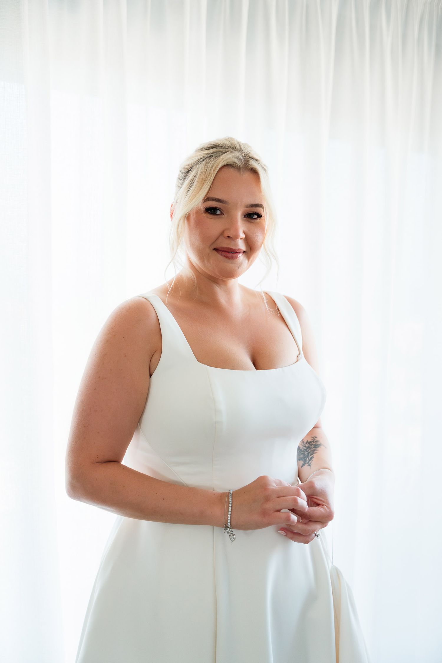 Bride in a white wedding dress gets ready against a bright window backdrop with soft white curtains.