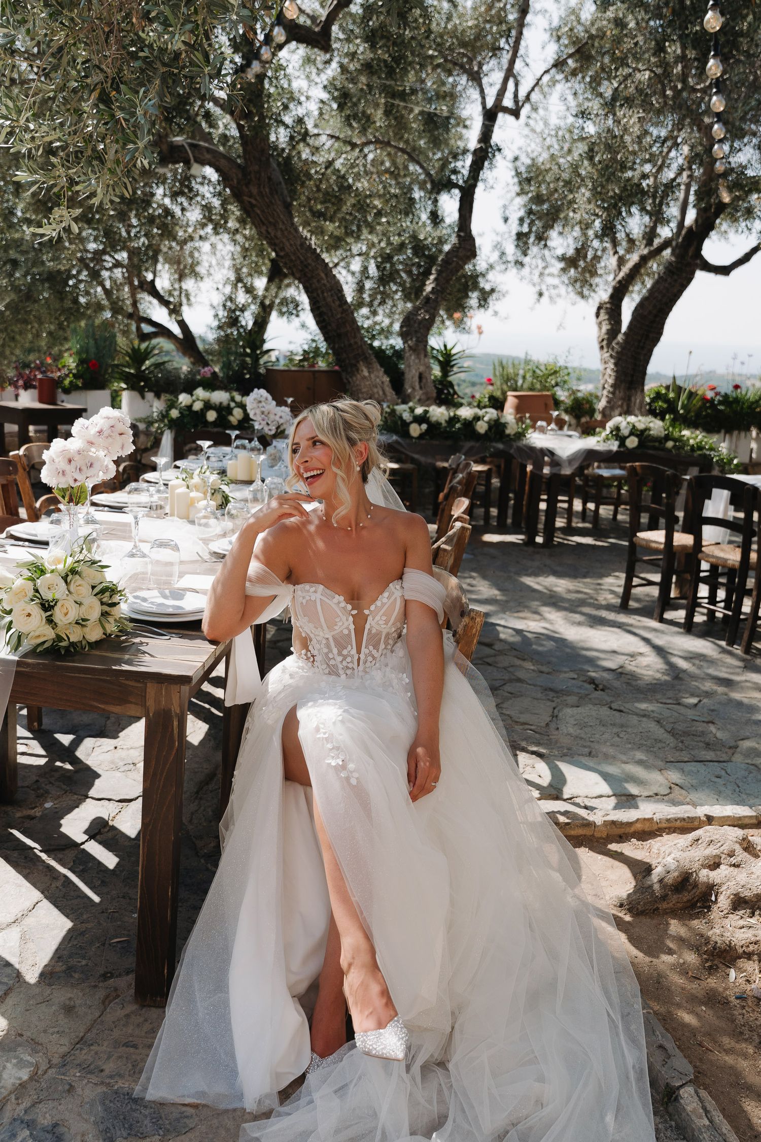 Elegant outdoor wedding reception setup under olive trees with rustic wooden tables and white floral arrangements.