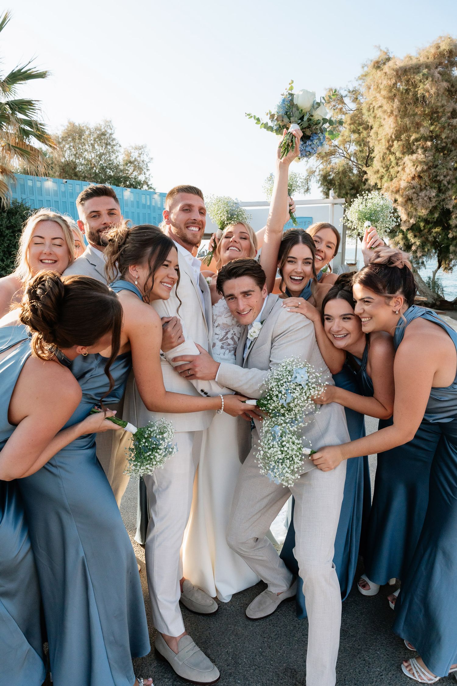 Wedding party in dusty blue bridesmaid dresses and light suits share joyful group hugs outdoors on sunny day.