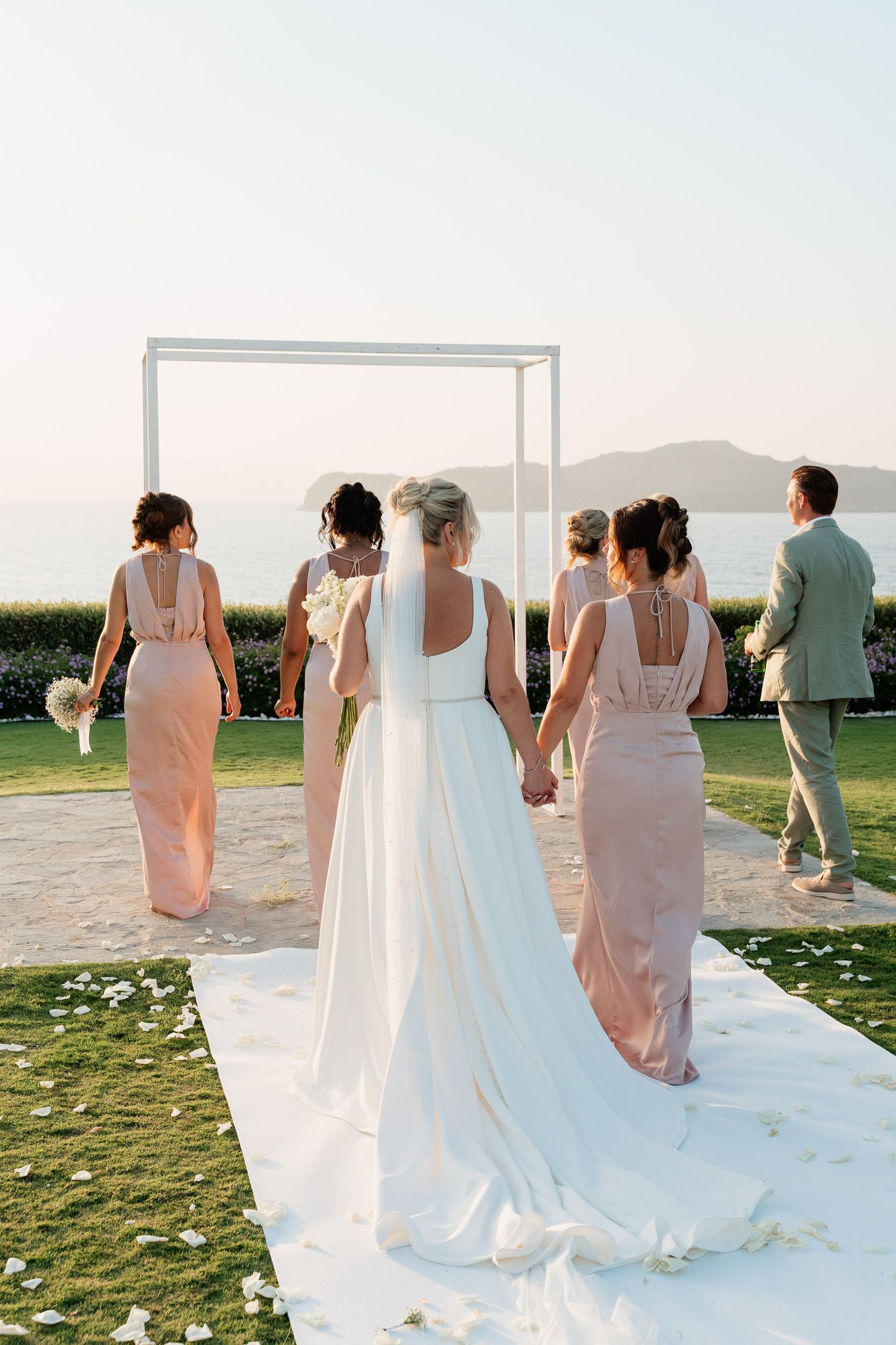 A wedding party in blush pink dresses and a flowing white gown stand at an outdoor ceremony overlooking coastal mountains.