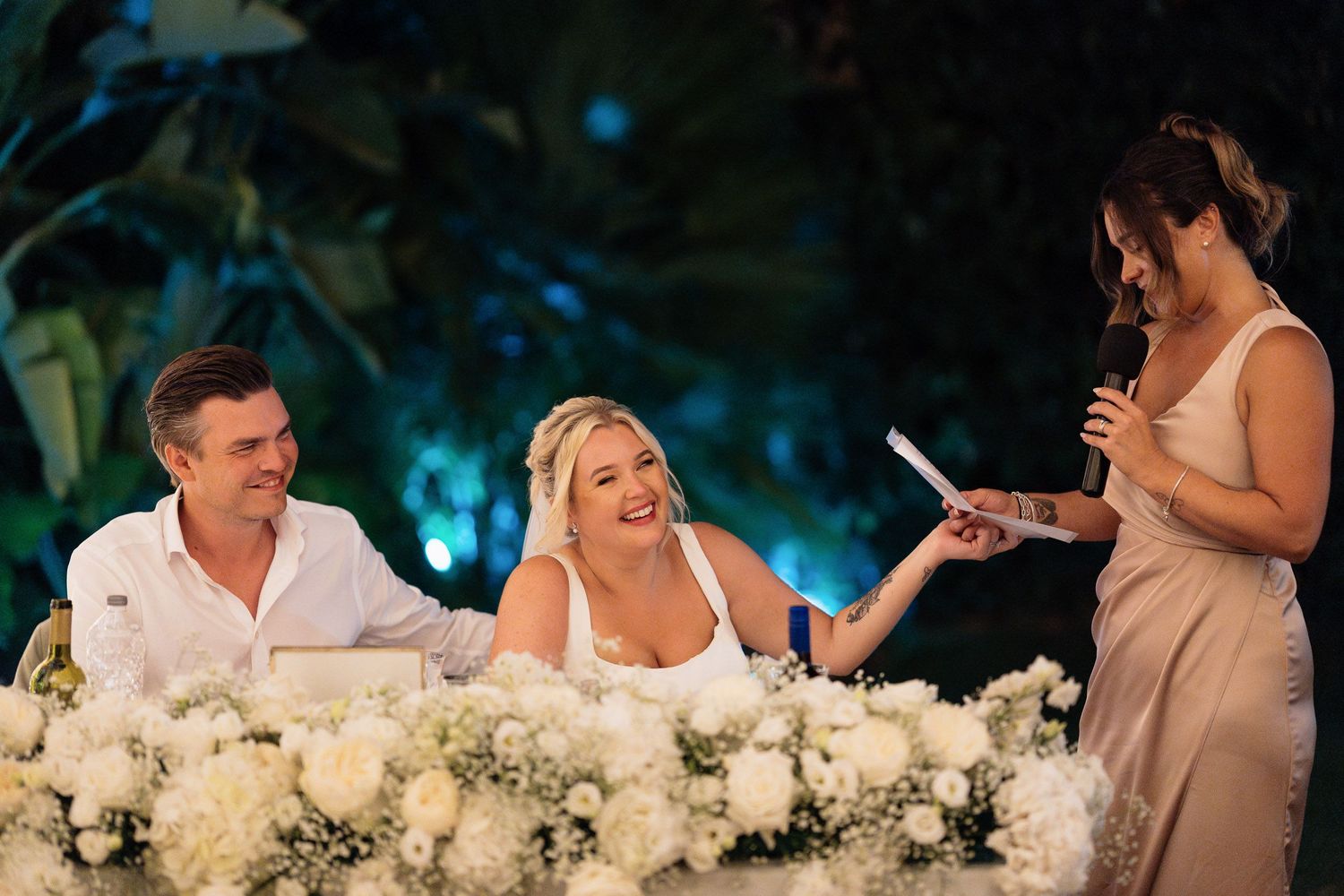 Wedding reception scene at an outdoor venue with people seated at a flower-adorned table during evening speeches.