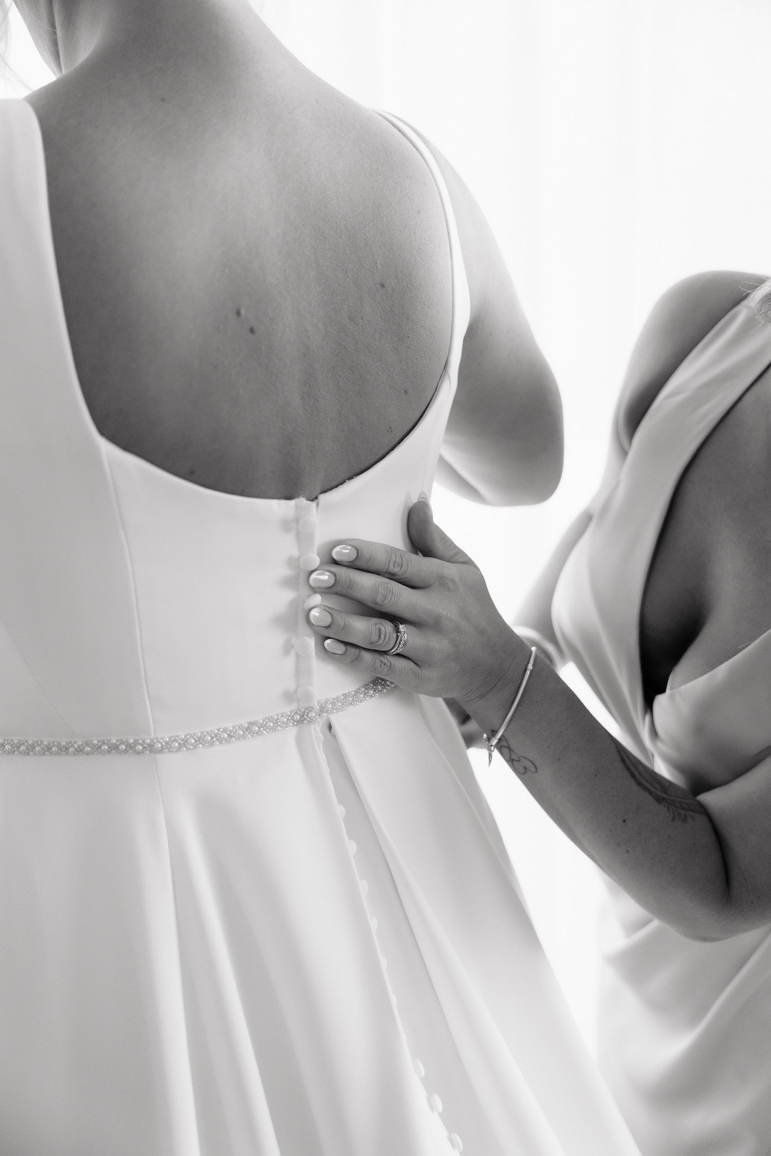 A black and white photo showing someone helping to button up the back of a white wedding dress.