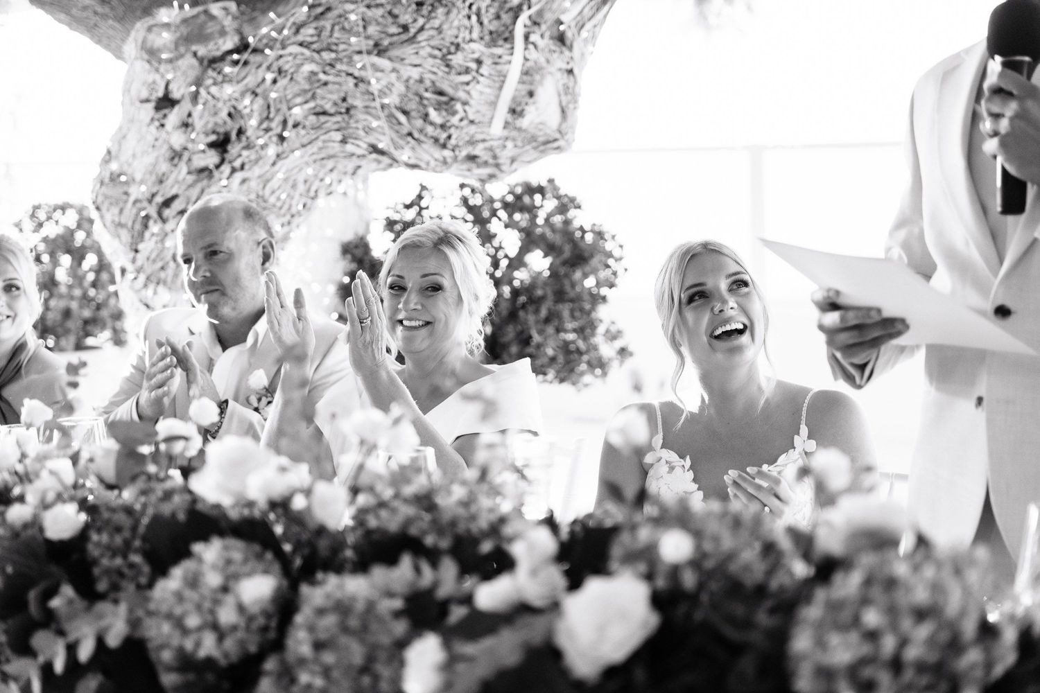 Black and white photo showing guests laughing and celebrating at an outdoor event with floral decorations.