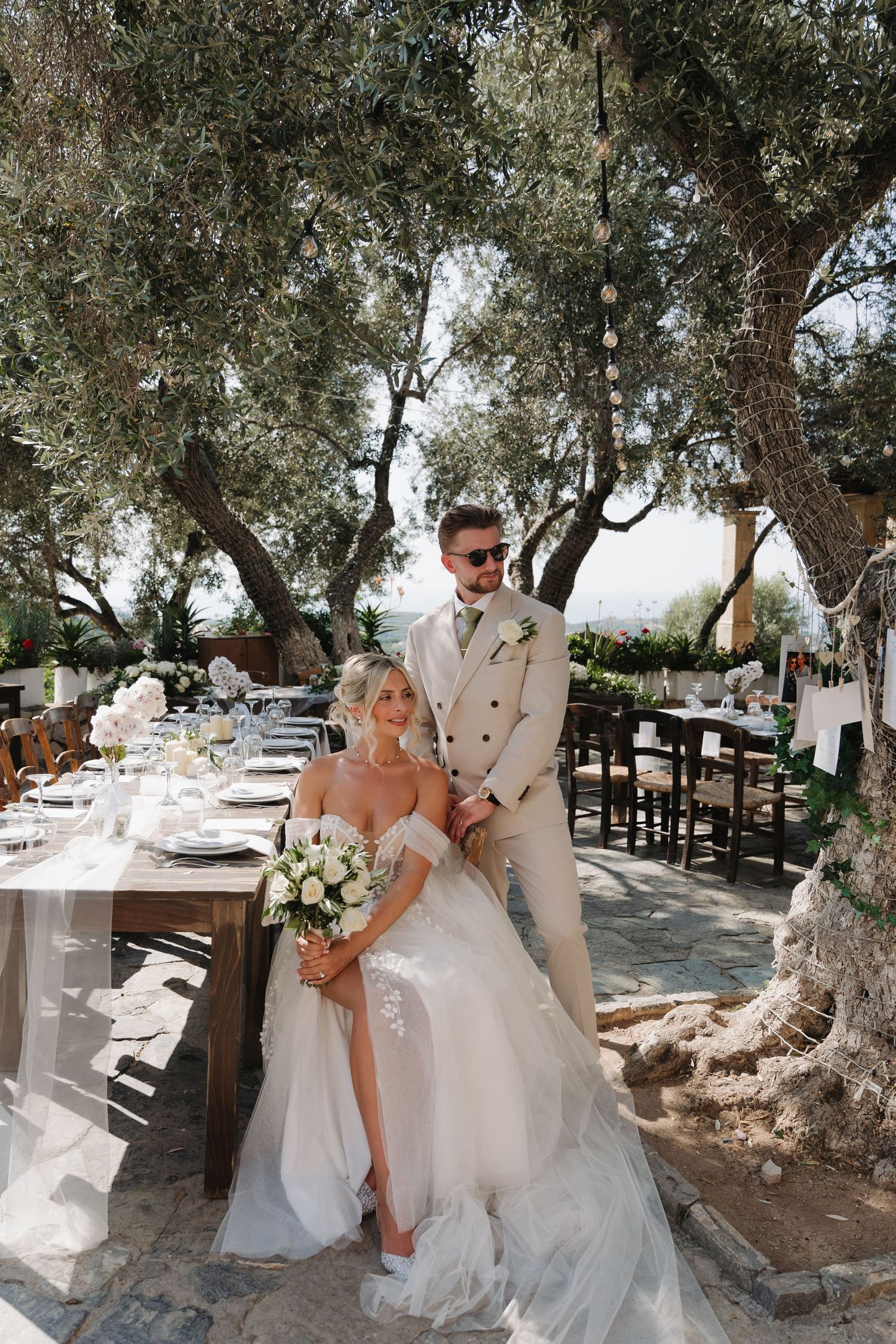 Elegant outdoor wedding ceremony setup under olive trees in a Mediterranean coastal setting with white decor and natural scenery.