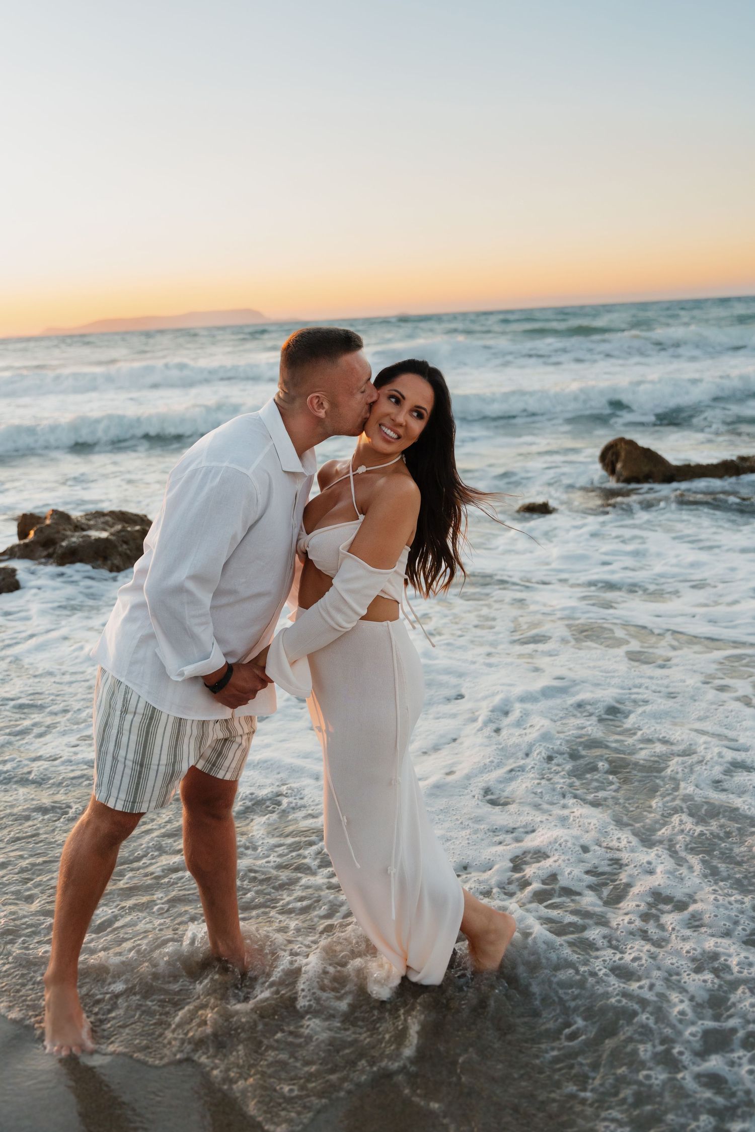 A couple shares a romantic moment on a beach at sunset as waves crash around their feet.