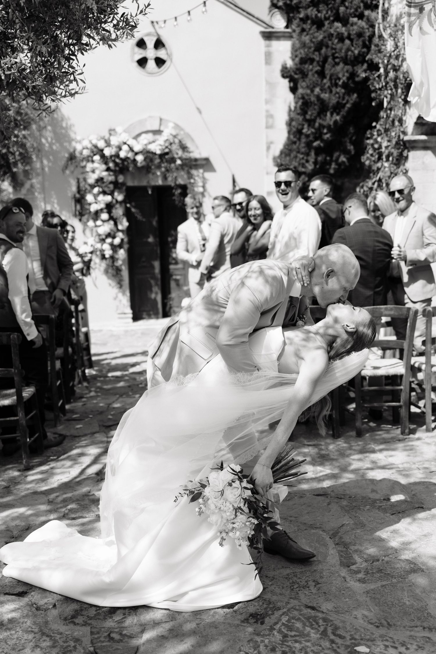 Groom dips bride for a romantic kiss at outdoor wedding ceremony in black and white photography.