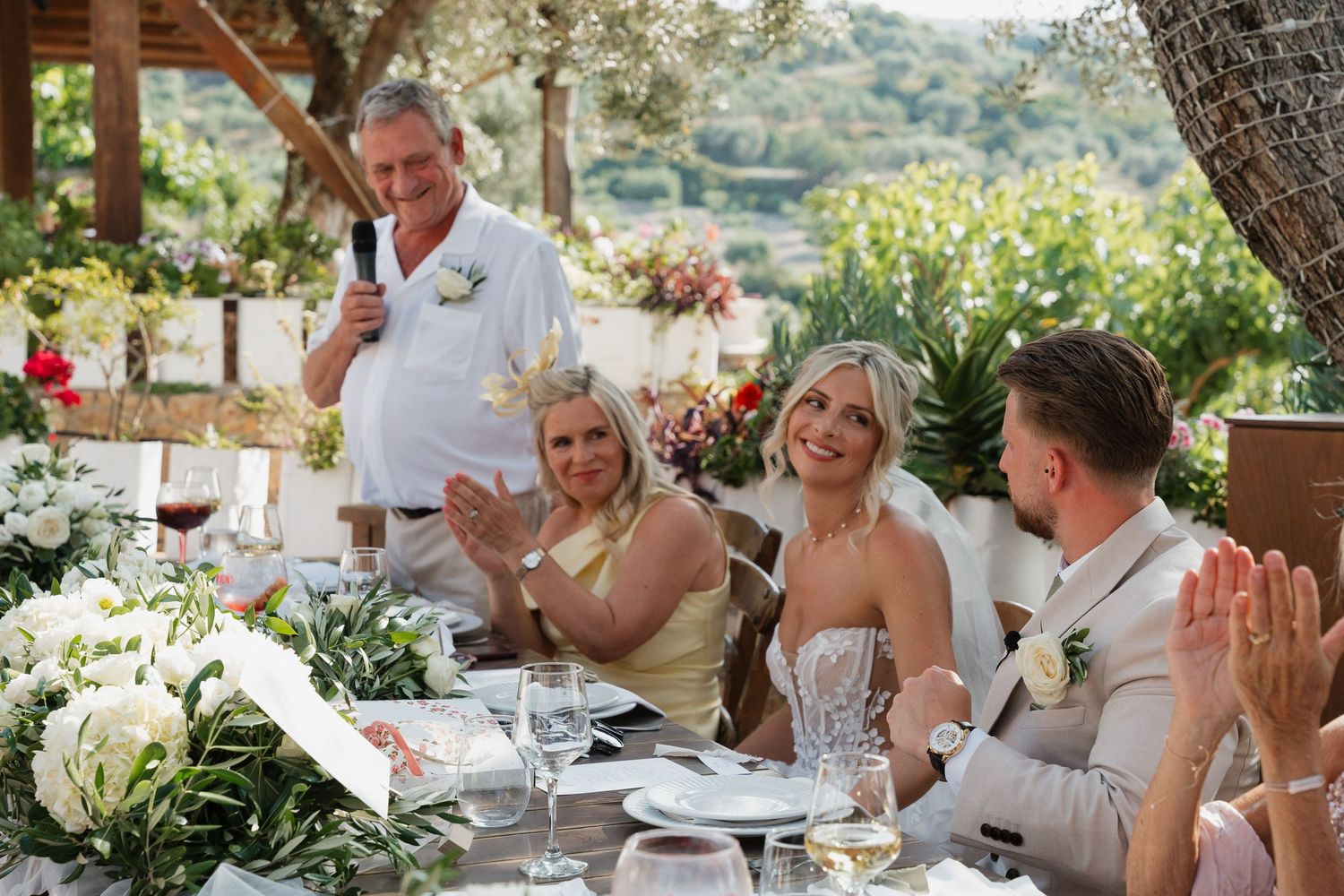 Wedding guests enjoy an elegant outdoor reception dinner under a covered patio surrounded by lush greenery and flowers.