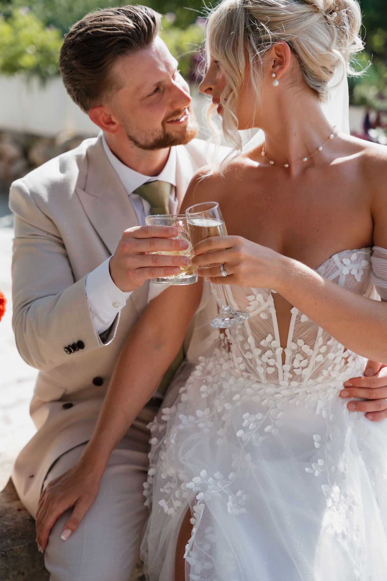 A bride in a white lace gown and groom in beige suit share a toast while smiling at each other on their wedding day.