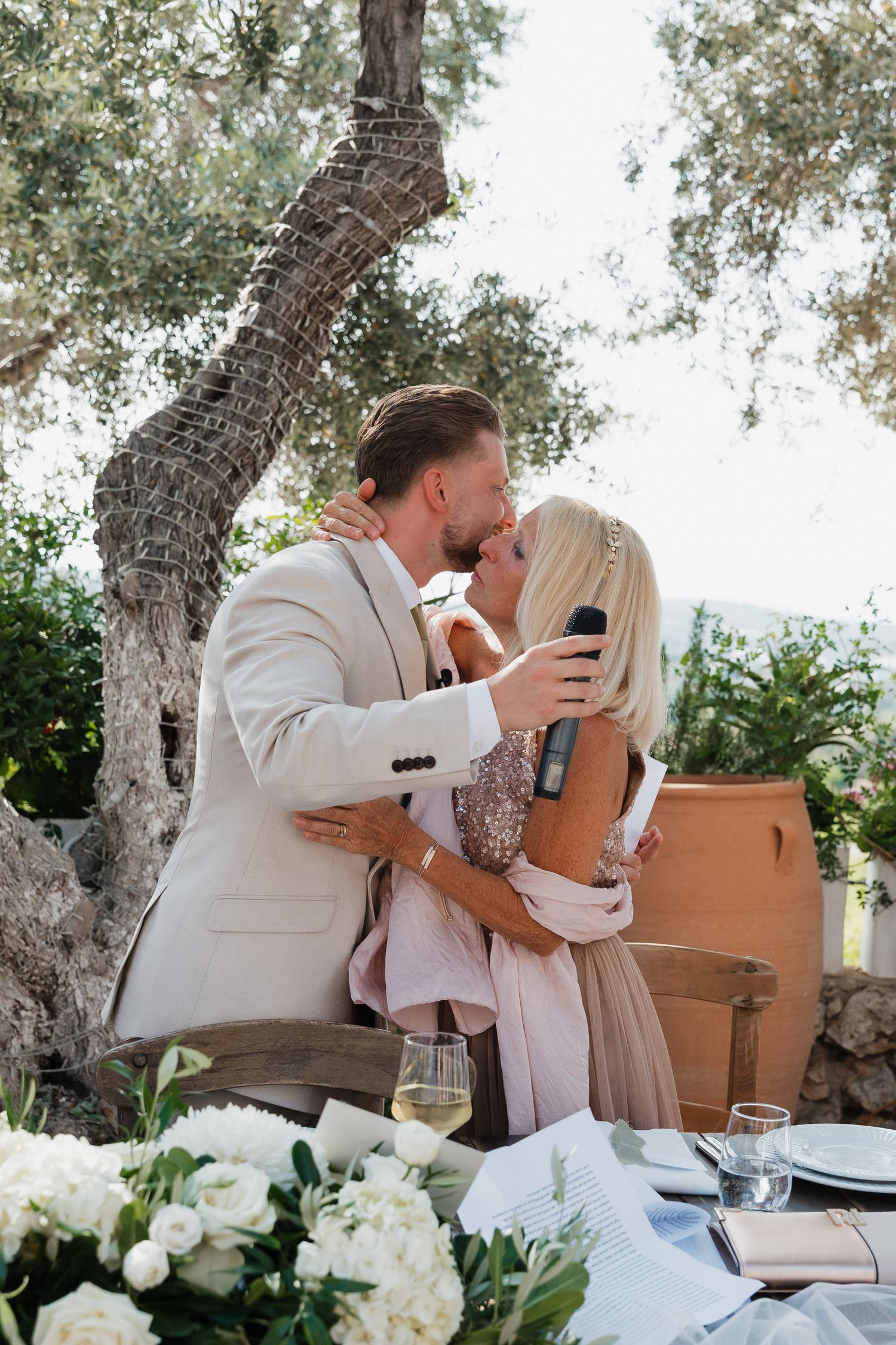 A romantic outdoor dining scene with white floral arrangements and wine glasses under a twisted olive tree.