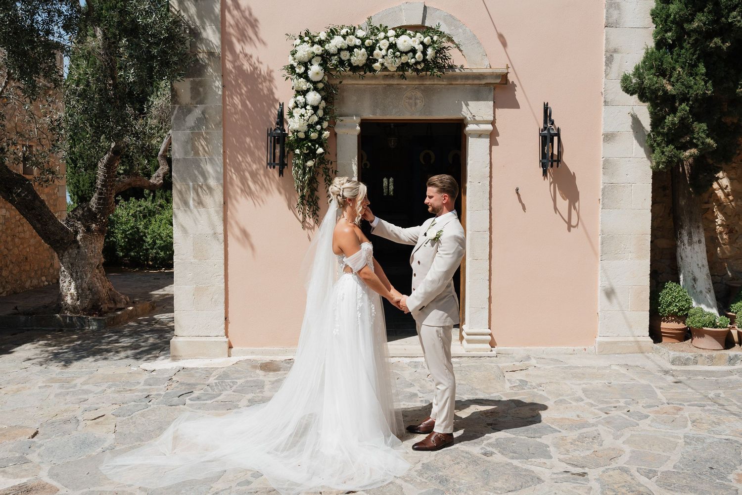 A bride in a flowing white wedding dress walks along a rustic pink stone wall in an elegant outdoor setting.
