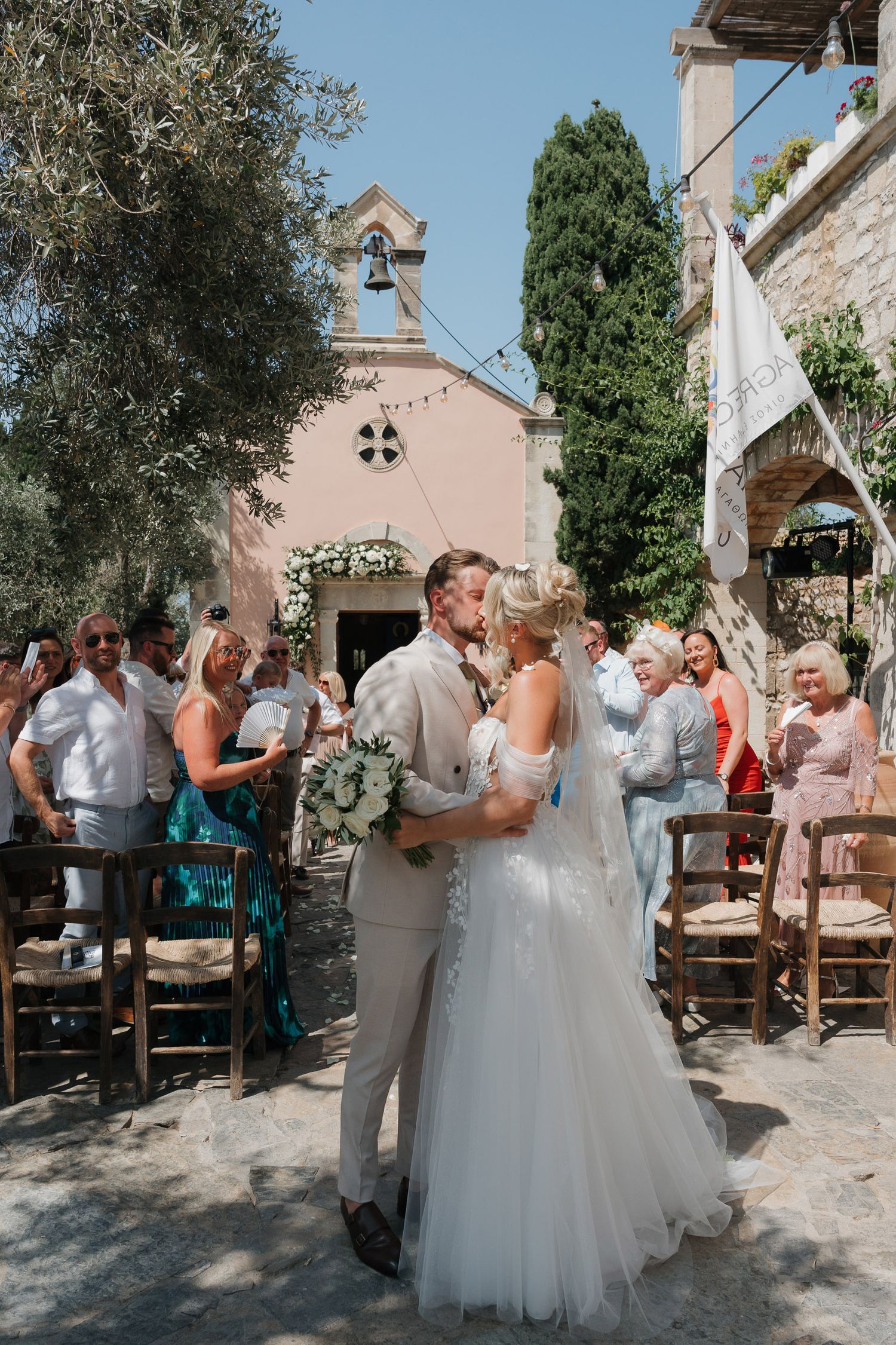 A wedding ceremony takes place outside a charming pink chapel with a bell tower in a Mediterranean setting.