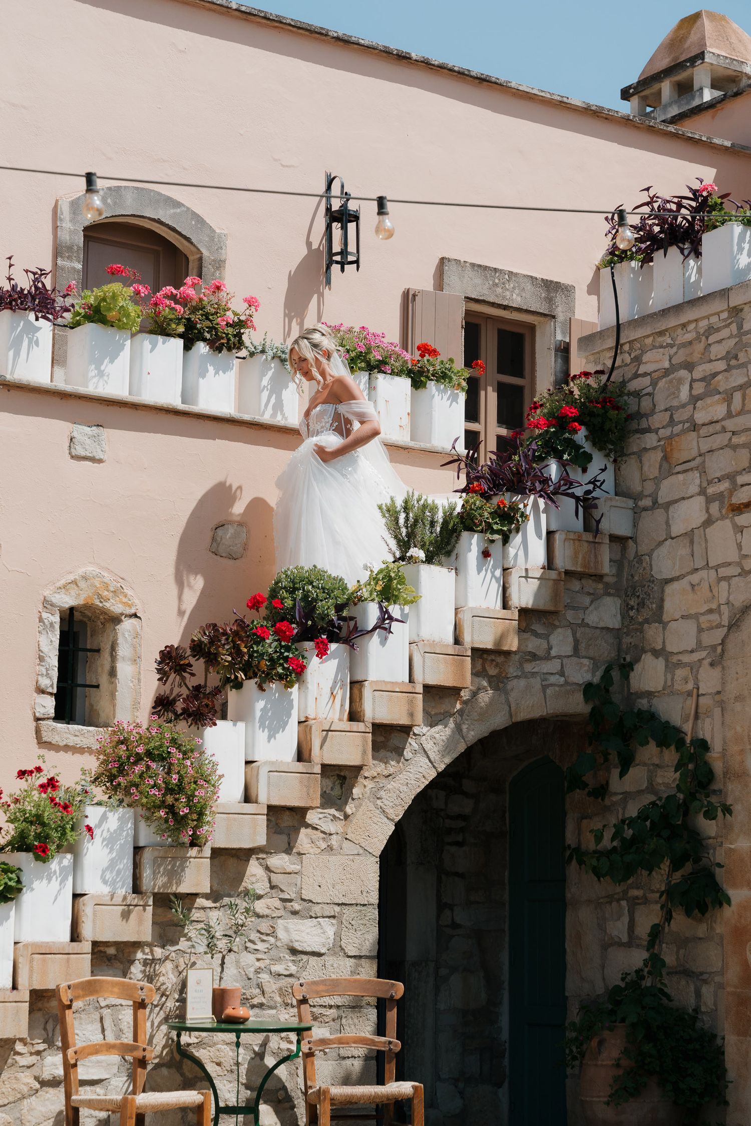 Stone archways and steps adorned with pink flowers and white curtains create a charming Mediterranean courtyard atmosphere.