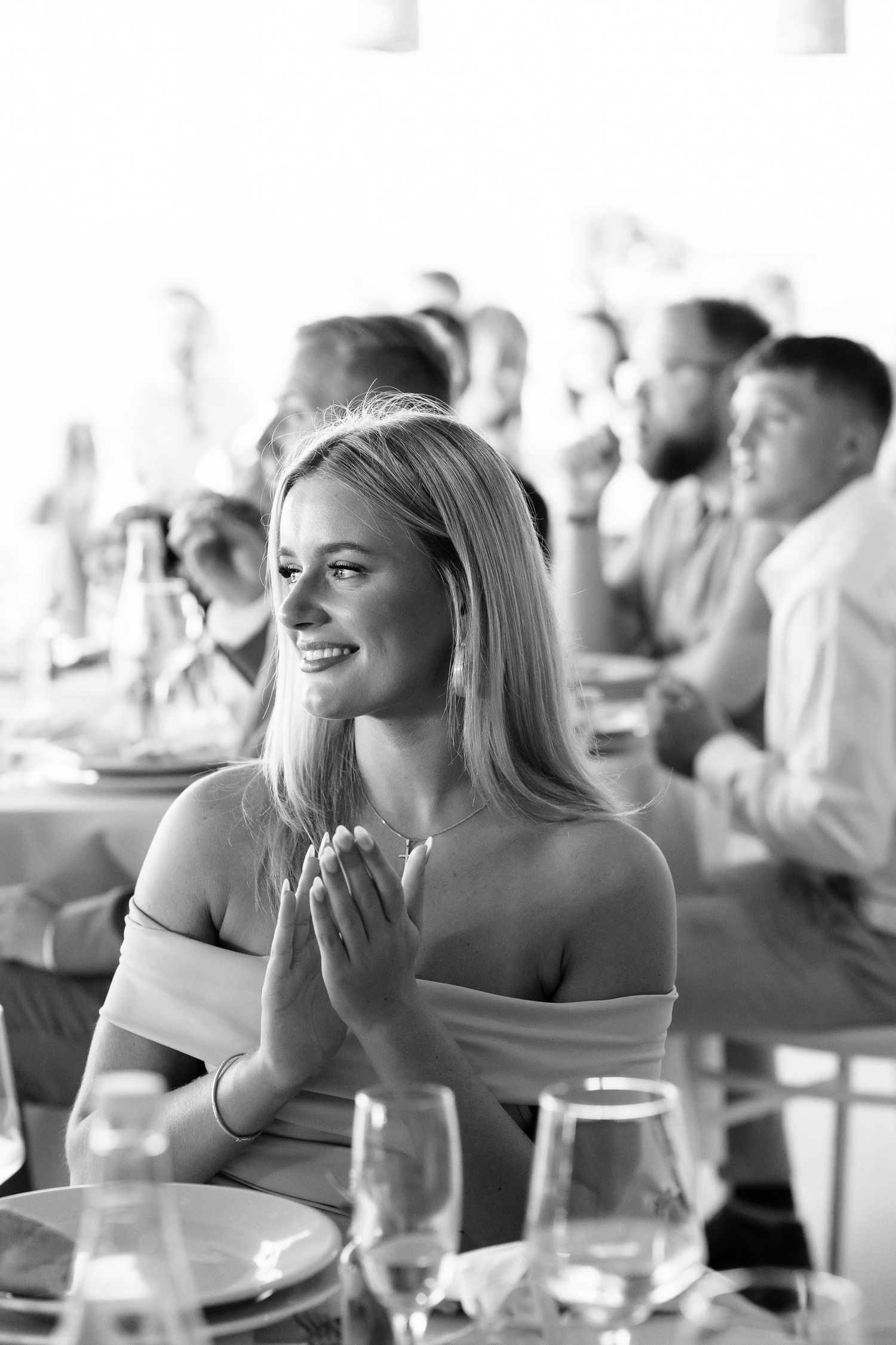 A woman in an off-shoulder dress smiles brightly during a formal dining event with guests in the background.
