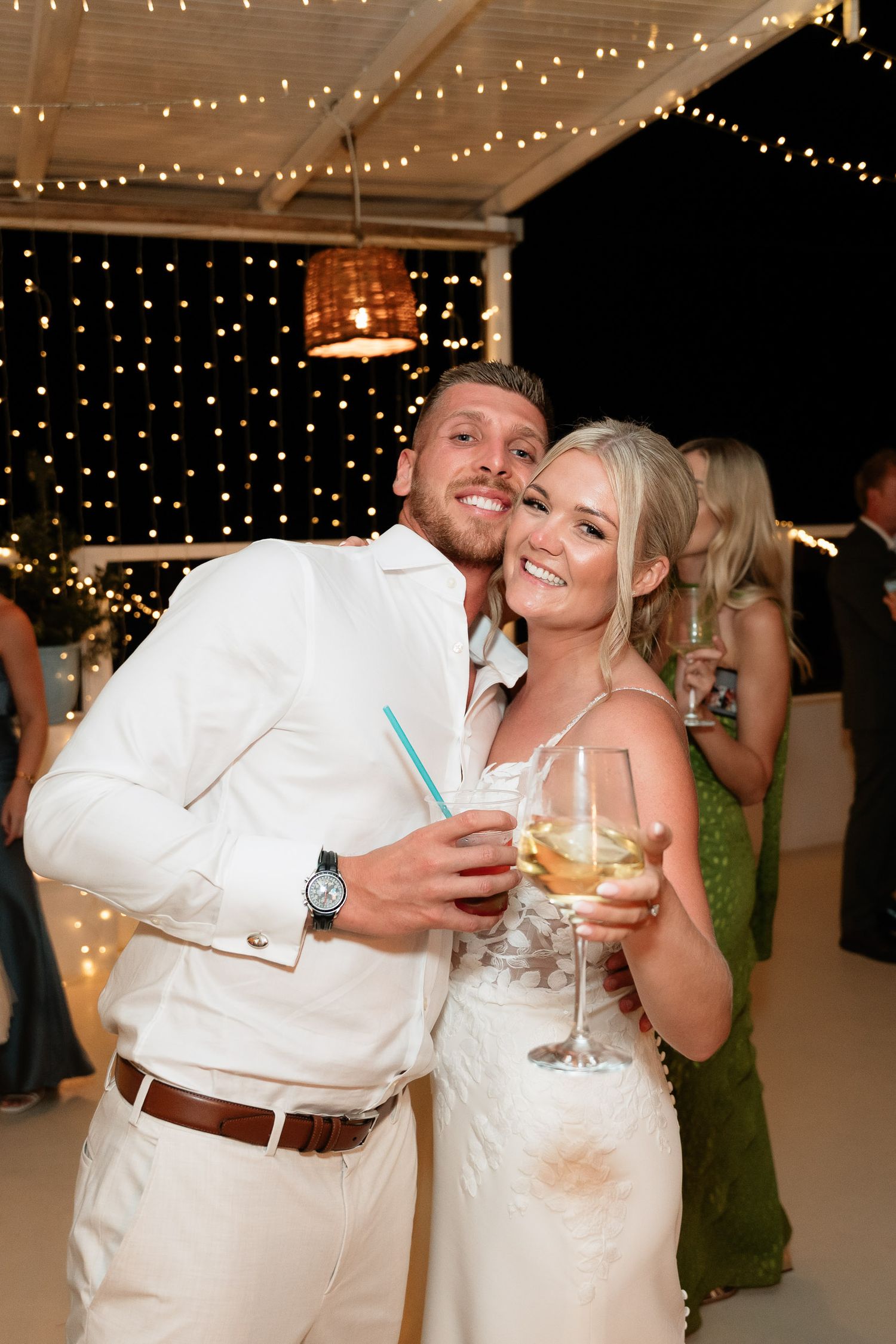 A couple celebrates at a wedding reception under twinkling string lights while holding cocktails.