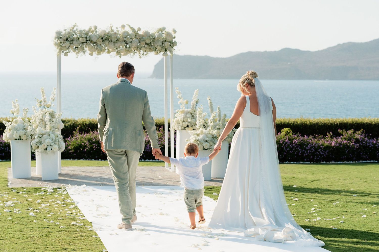 A wedding party walks along a scenic oceanfront lawn with mountains and white floral decorations in view.