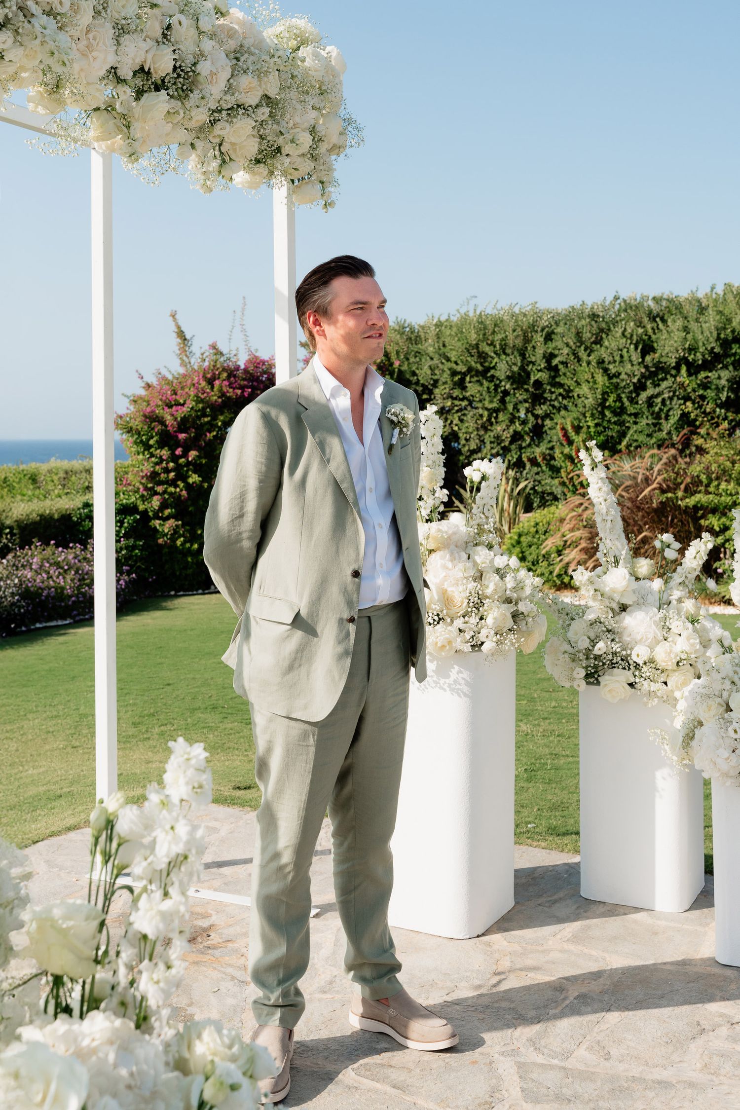 A person in a light beige suit poses next to elegant white floral arrangements at an outdoor seaside venue.