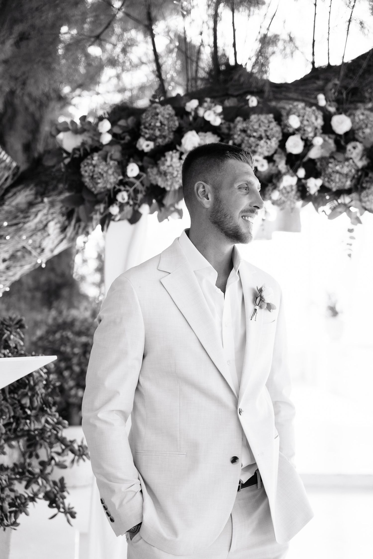 Black and white photo of a groom in a light suit standing under an elegant floral arch at a wedding venue.