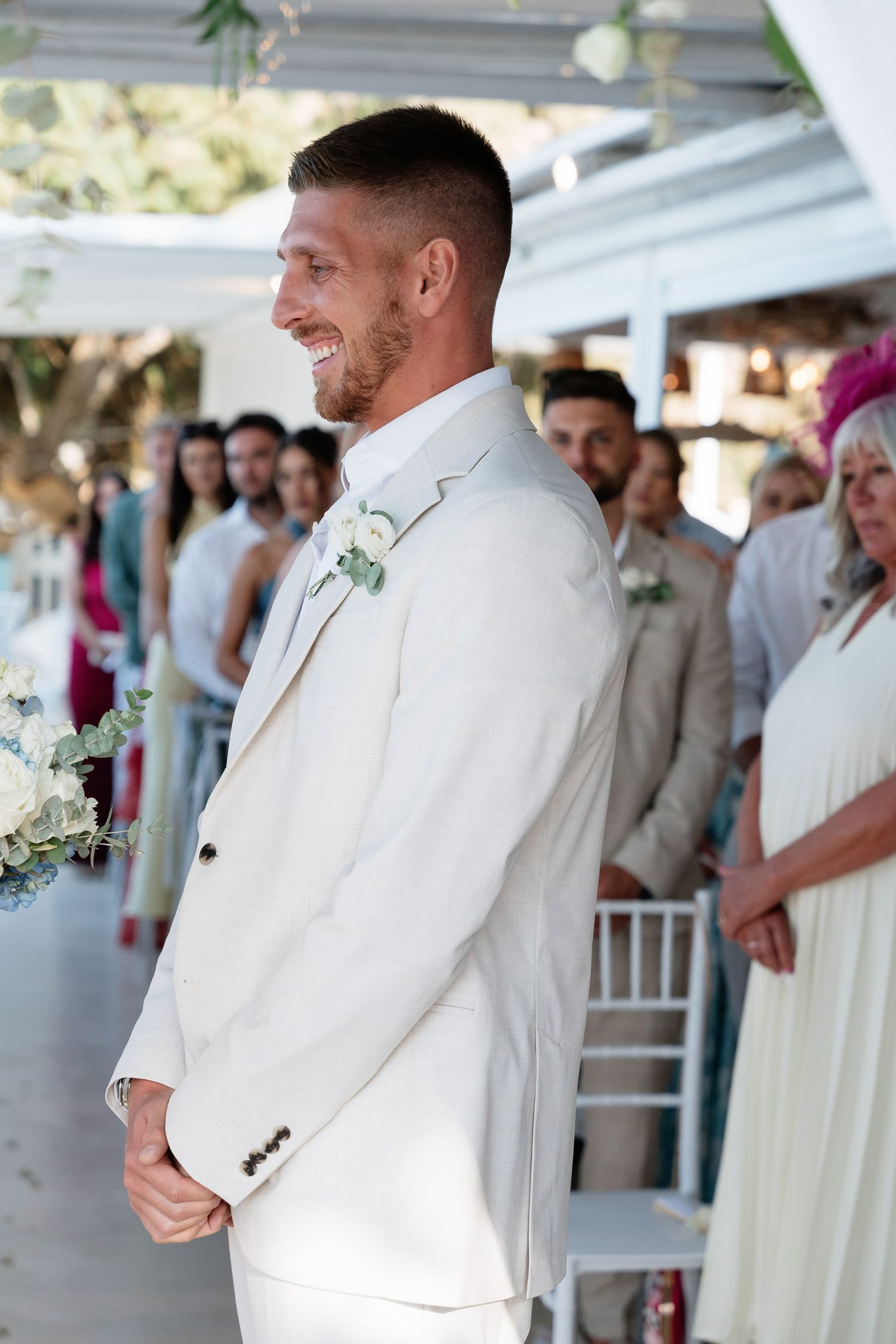 A groom in a white suit smiles while standing at an outdoor wedding venue with guests in the background.