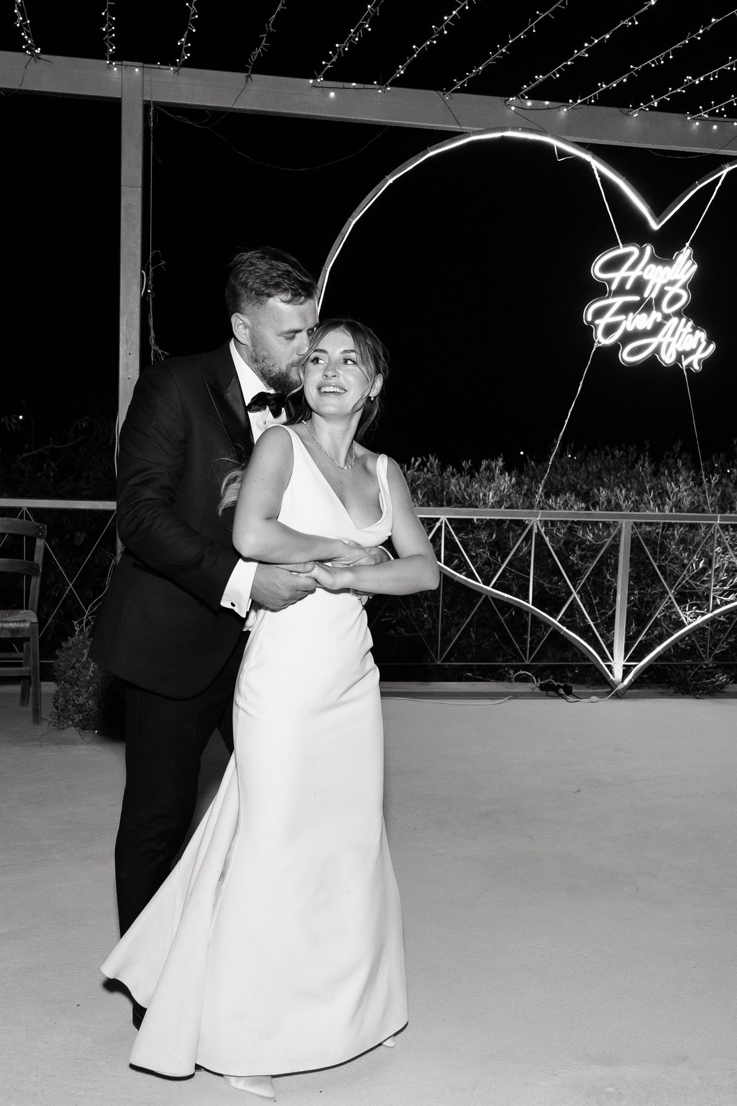 A black and white wedding photo showing a couple embracing on a patio with decorative lights in the background.