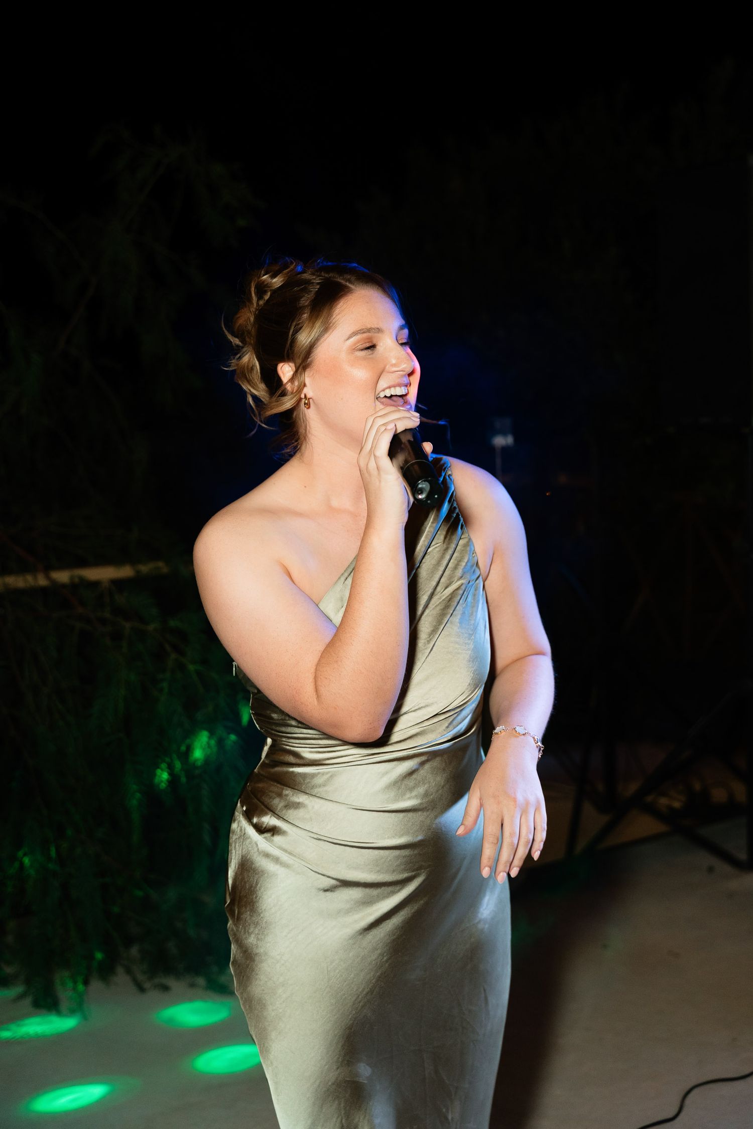 A person in a light-colored dress laughs and dances expressively during an evening event with green lighting.