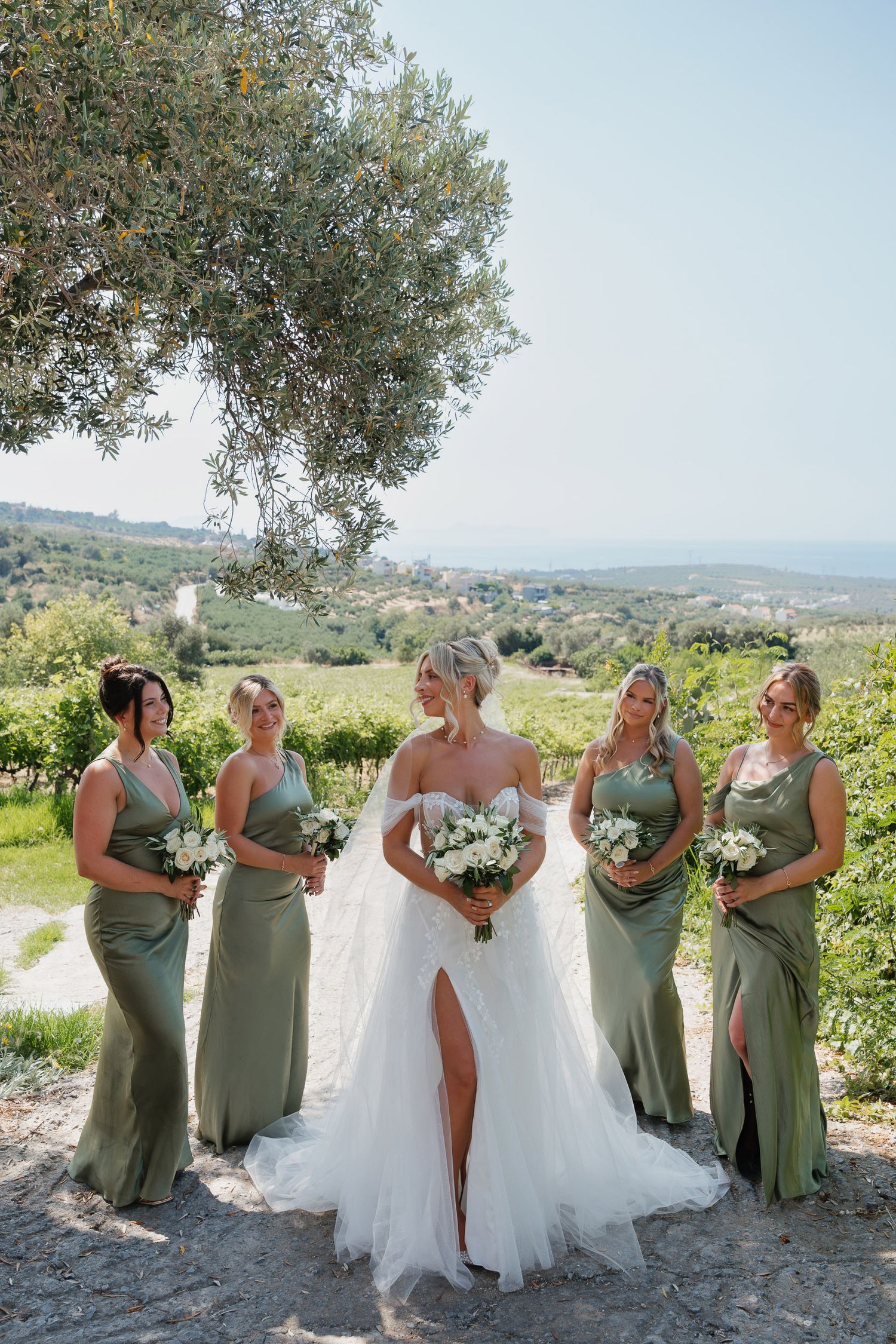 A bridal party in sage green dresses poses together against a scenic Tuscan hillside landscape under an olive tree.