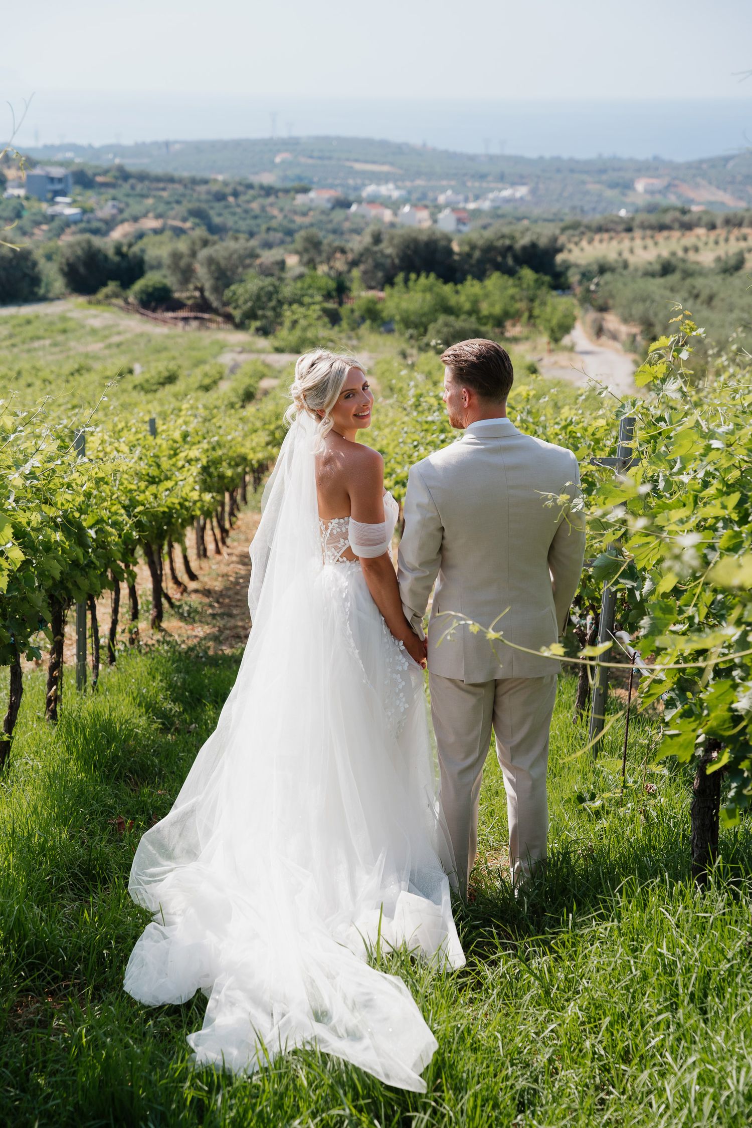 A couple in wedding attire stands together overlooking a scenic vineyard landscape on a sunny day.