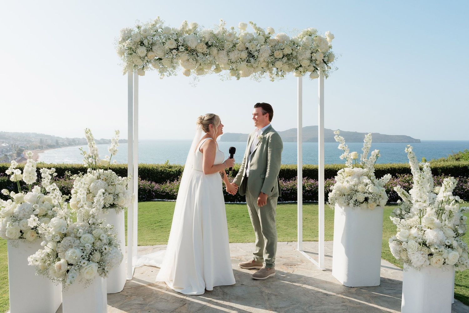 A wedding ceremony setup with white floral arrangements and columns against a scenic coastal backdrop.