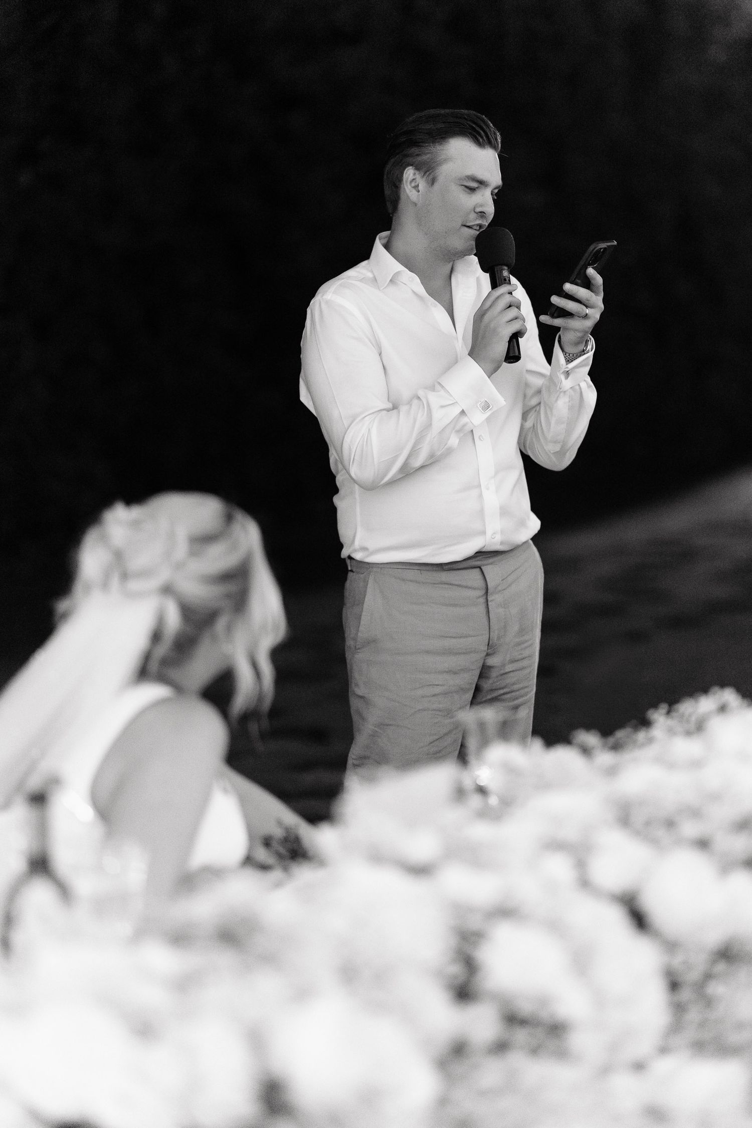 Black and white photo of a wedding moment showing someone checking their phone while standing near floral arrangements.
