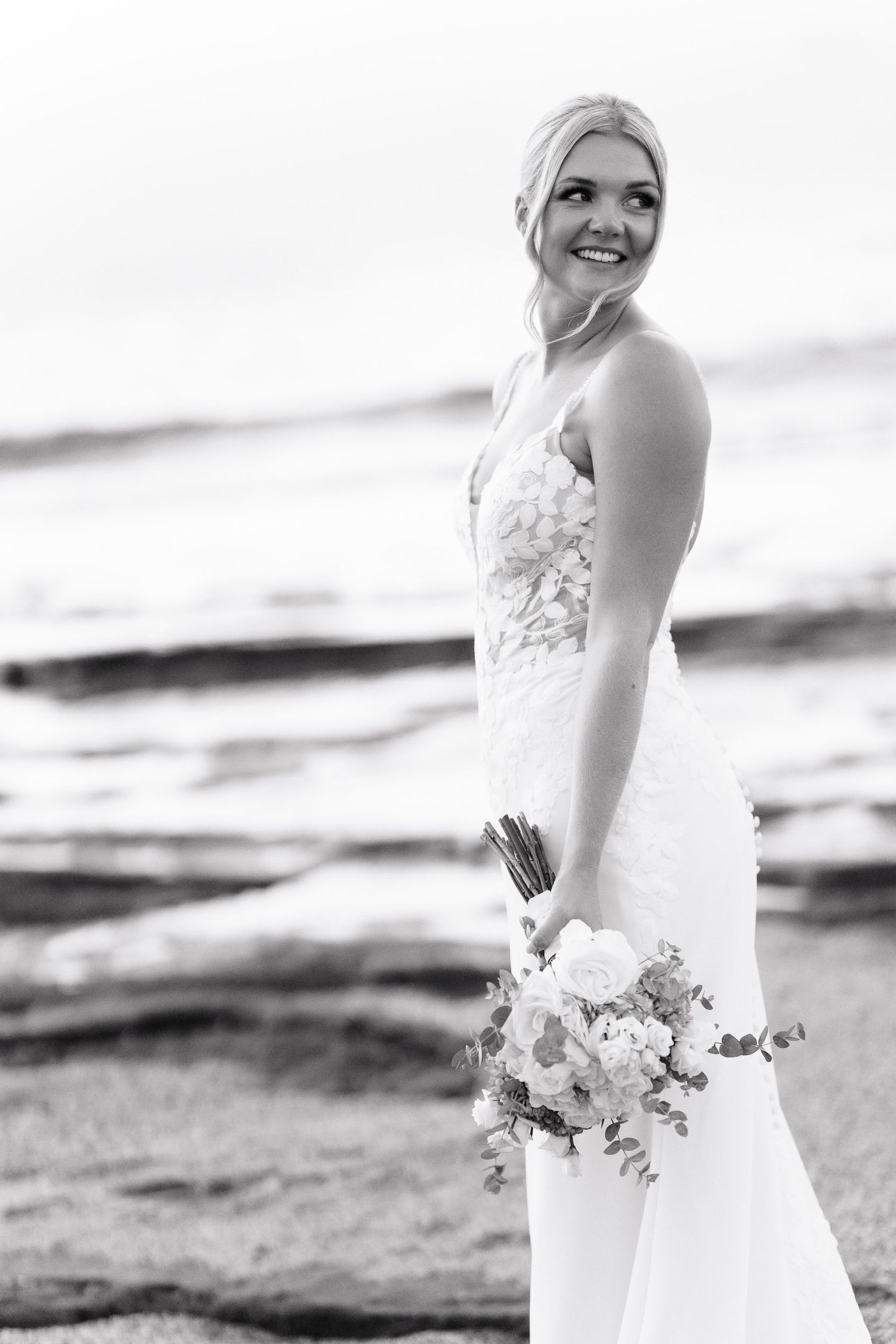 A bride in a white wedding dress holds a floral bouquet while posing on a beach at sunset.