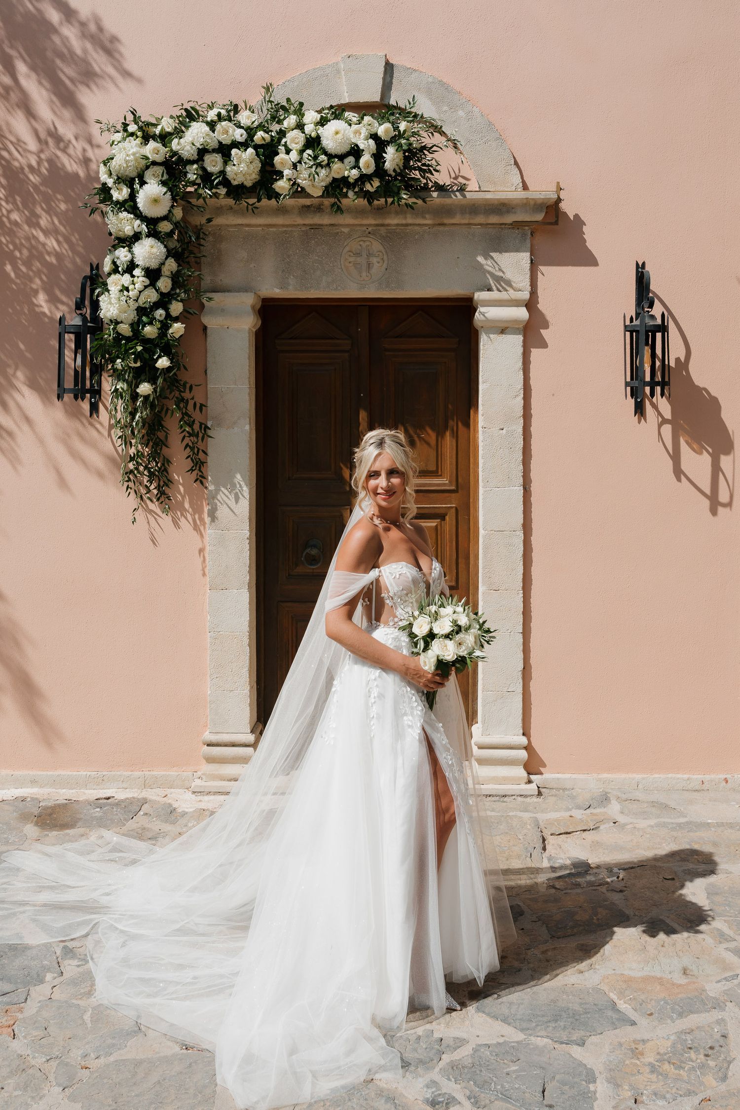 A flowing white wedding dress and veil create an elegant silhouette against a peach-colored Mediterranean style wall.
