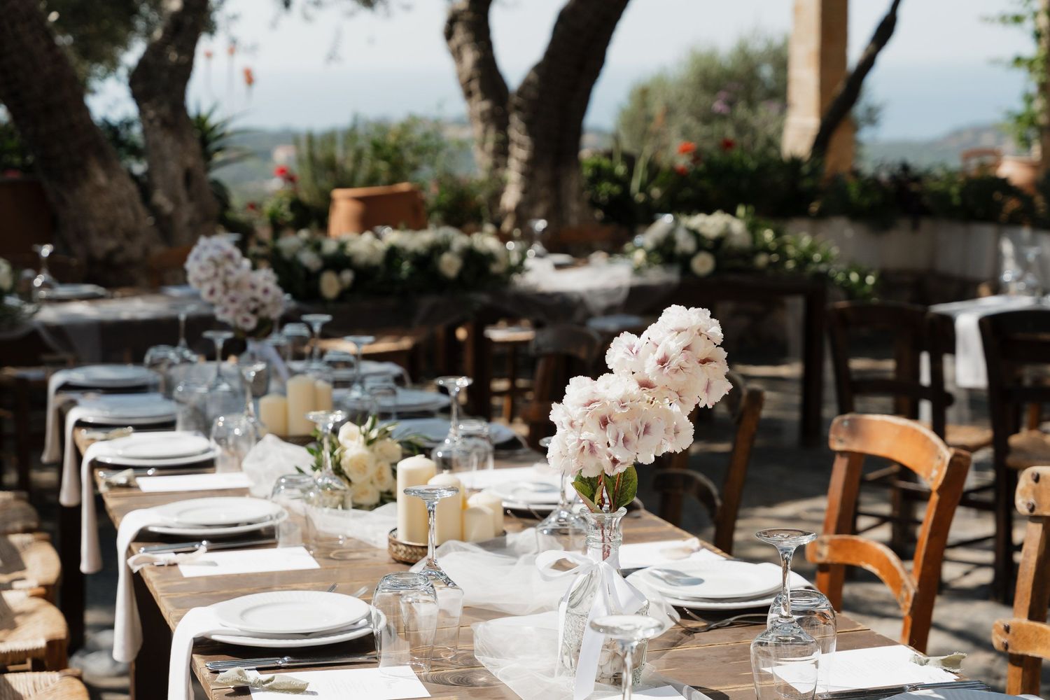 Elegant outdoor wedding reception table setting with pink floral centerpieces and rustic wooden chairs under trees.