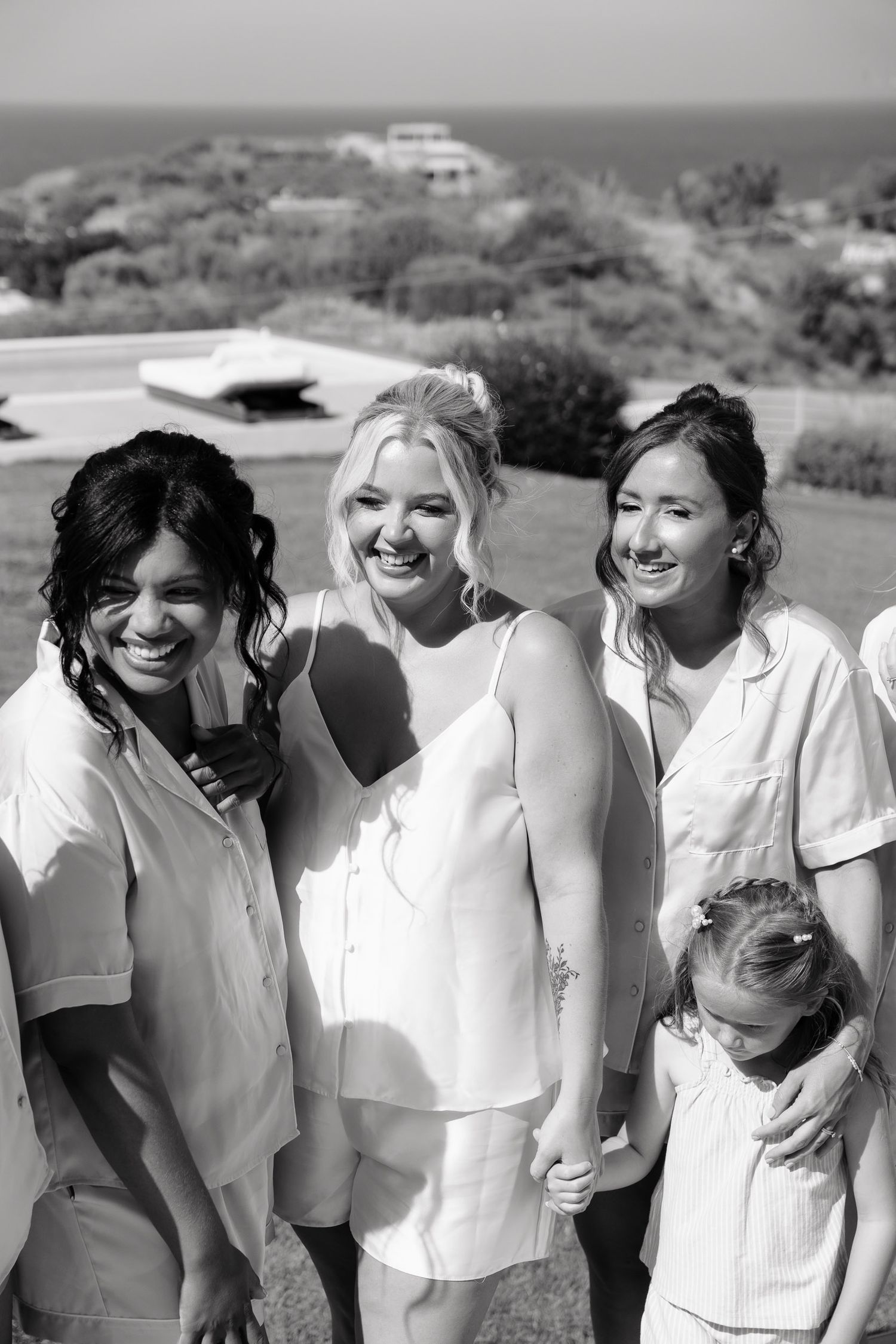 Black and white photo of a group in white outfits smiling together outdoors with ocean and buildings visible in background.