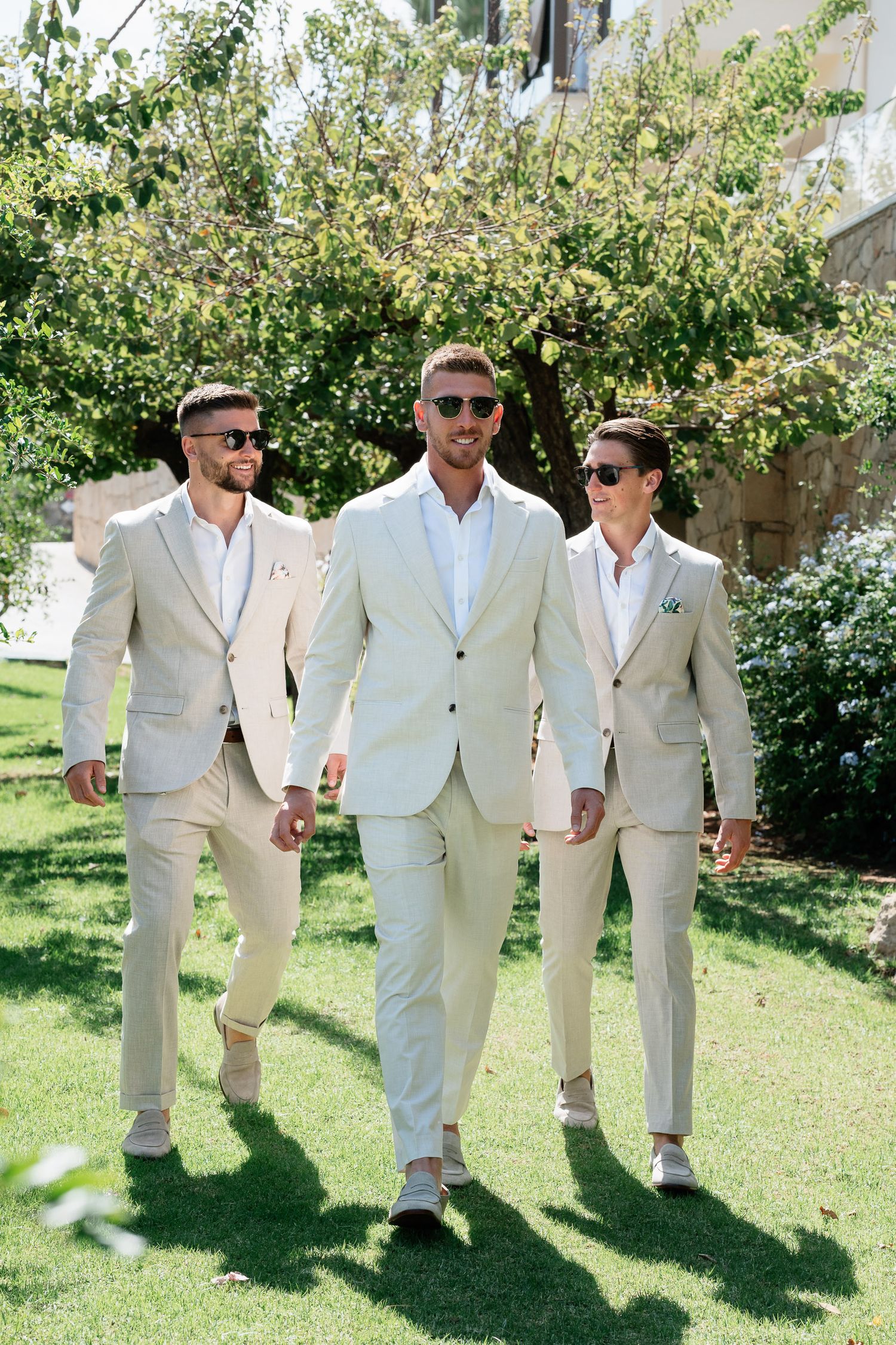 Three people in light colored suits and sunglasses walk together on a sunny lawn with greenery in the background.