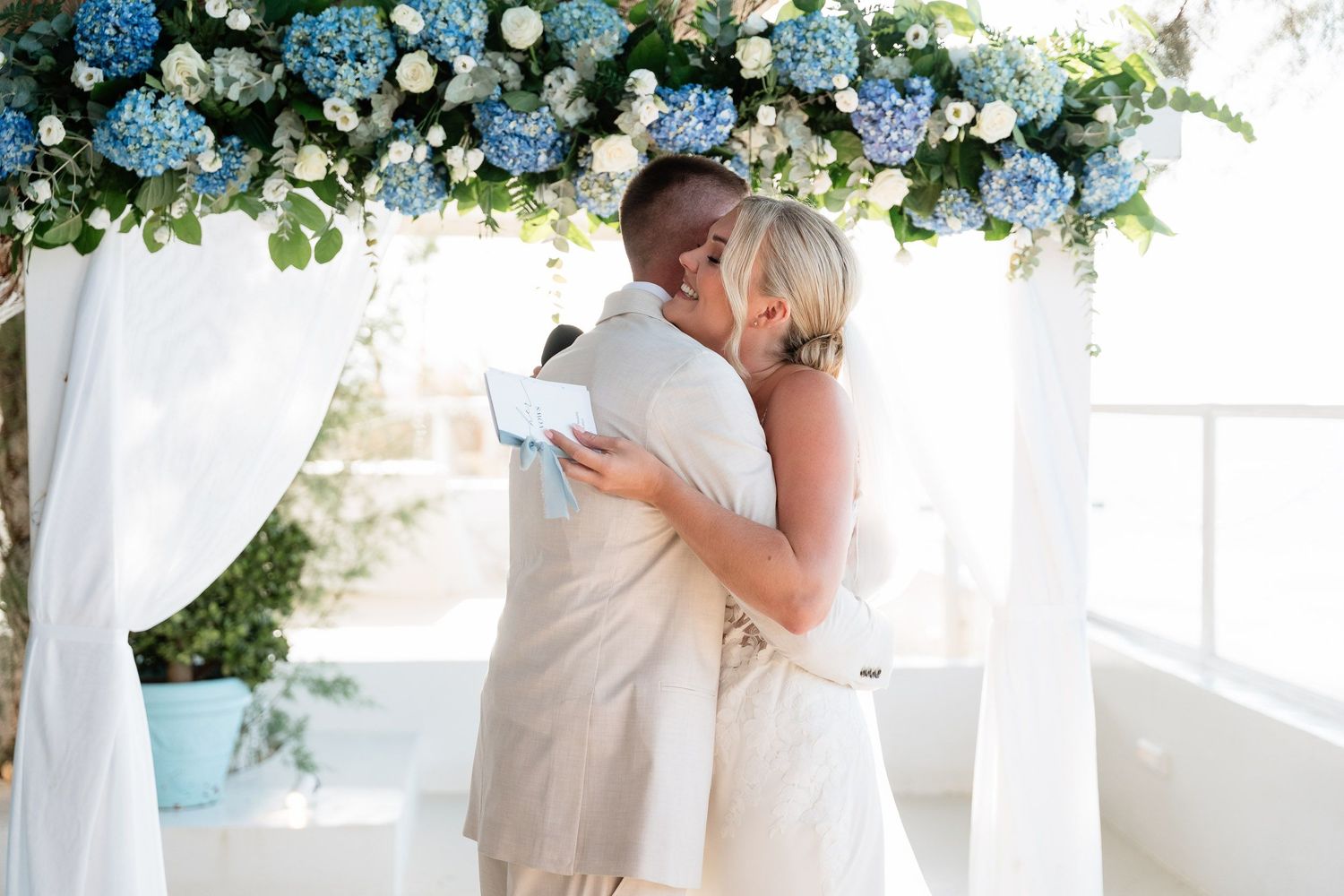 A couple shares a romantic kiss under an elegant floral arch with blue and white flowers at an outdoor wedding ceremony.
