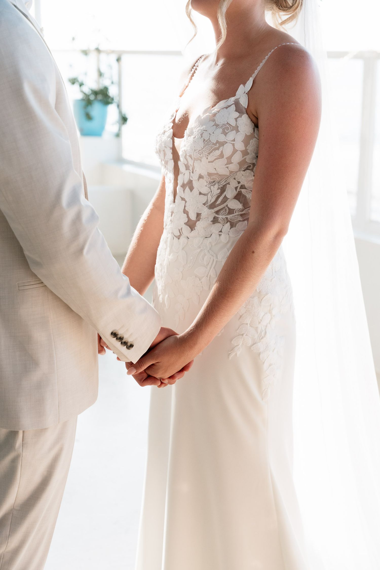 Close-up of couple holding hands at wedding, showing detailed lace bodice of white bridal gown and light colored suit.