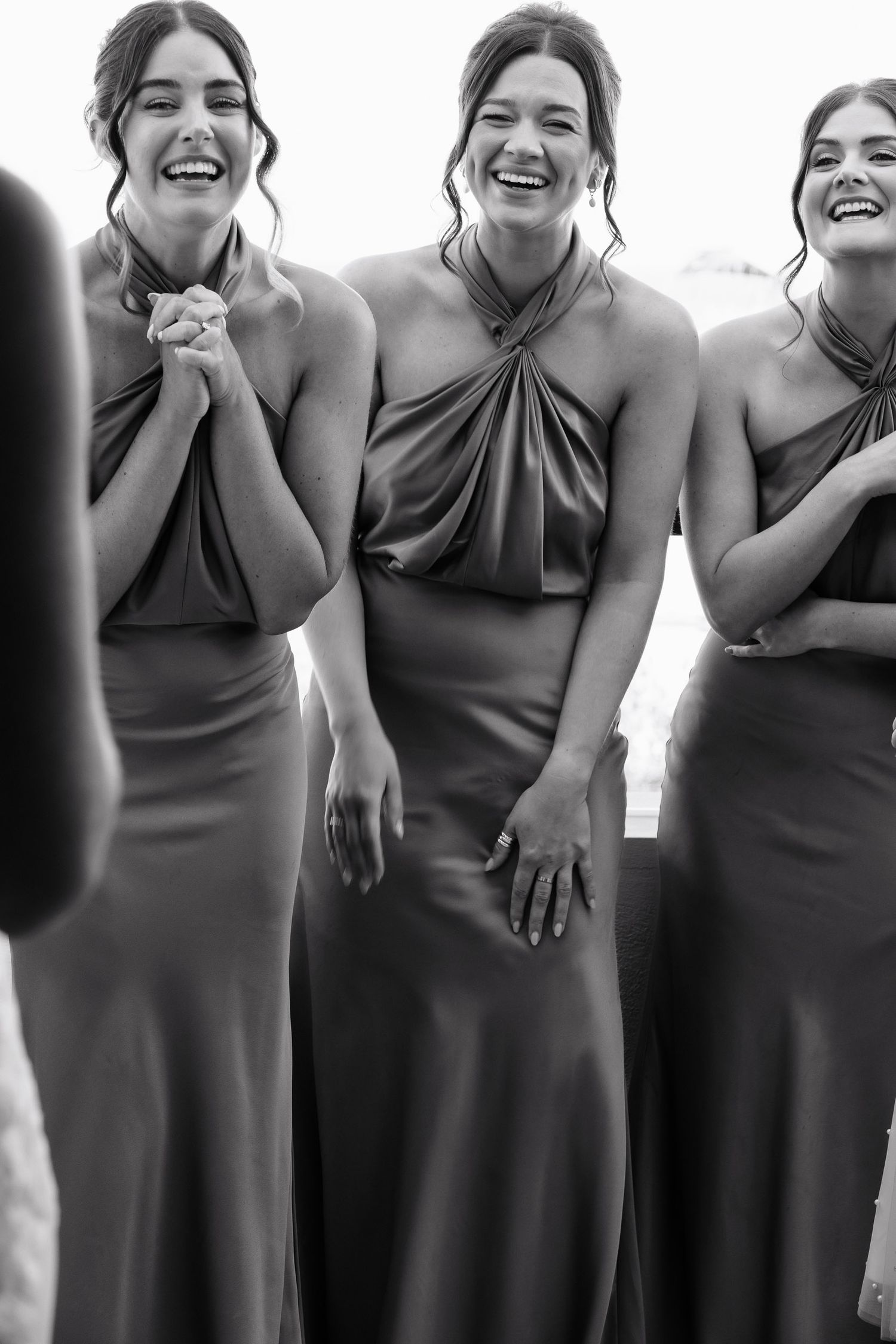 A group of bridesmaids in matching slate blue dresses share candid laughter in a black and white photo.