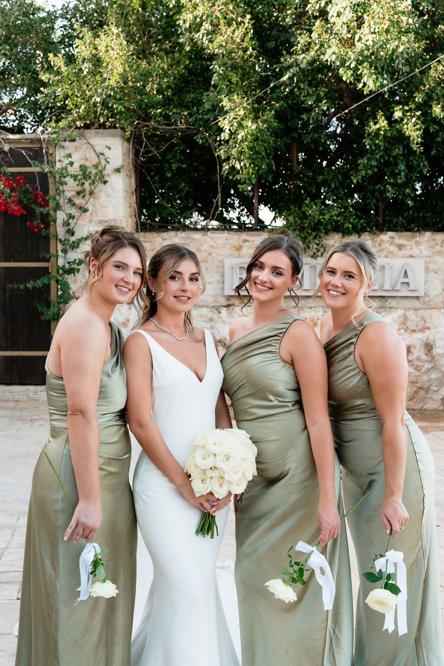 A bridal party in sage green dresses and white bouquets poses together outdoors against a stone wall in natural lighting.