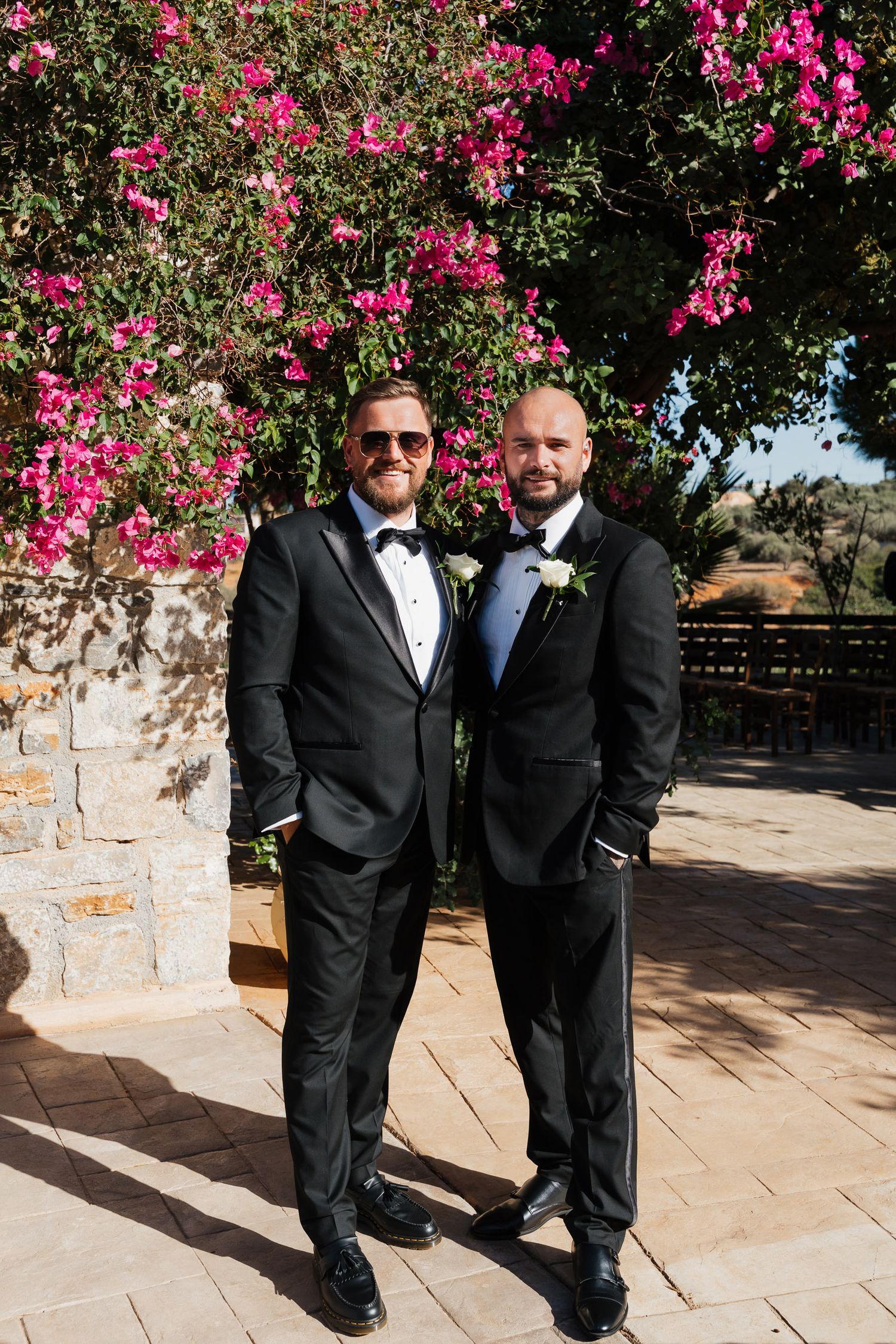 Two people in black tuxedos stand together under vibrant pink bougainvillea flowers against a stone wall.