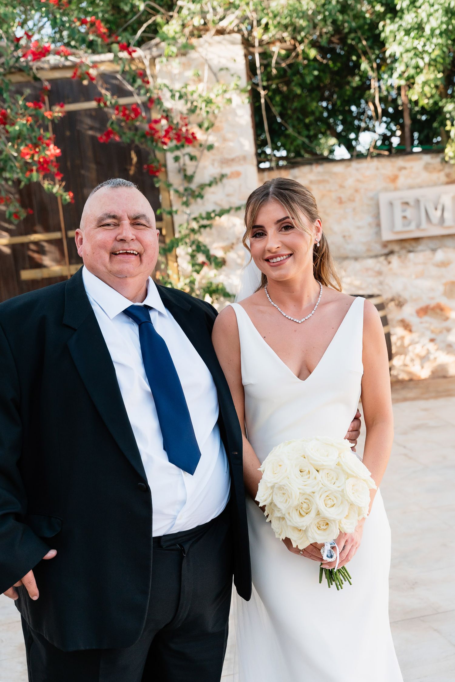 A bride in a white wedding dress poses with a companion outdoors under red flowers and greenery.