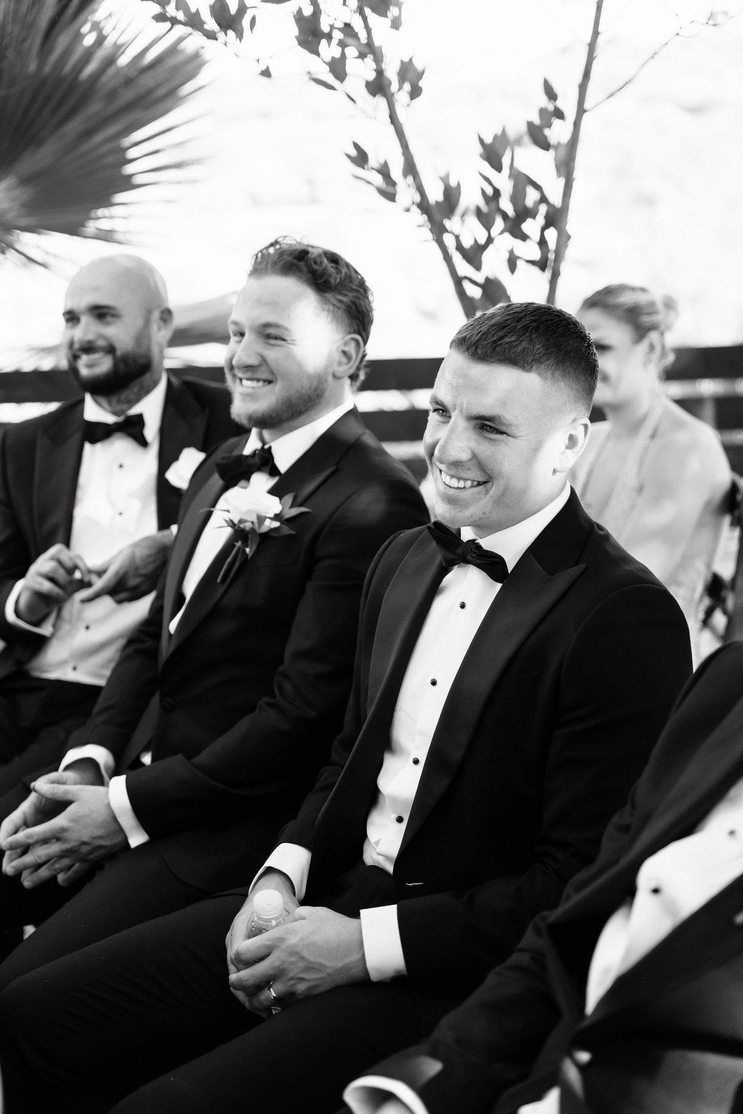 A black and white photo showing groomsmen in tuxedos laughing together during a wedding ceremony.