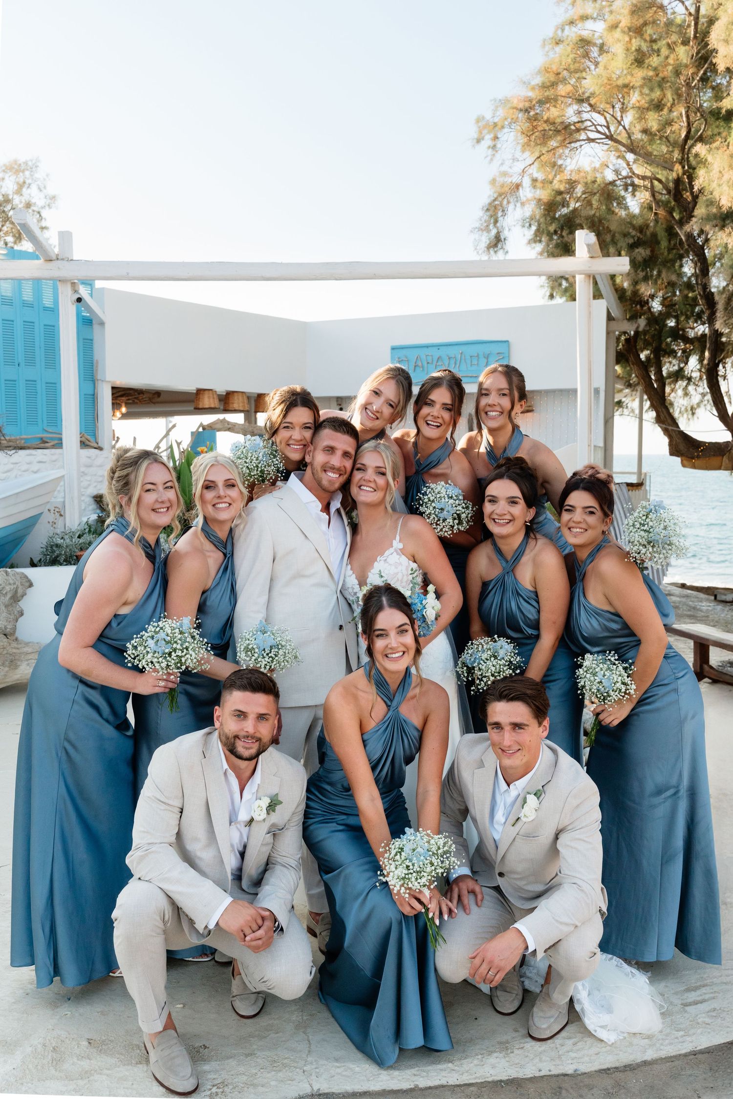 Wedding party in dusty blue bridesmaid dresses and light gray suits posing by waterfront.