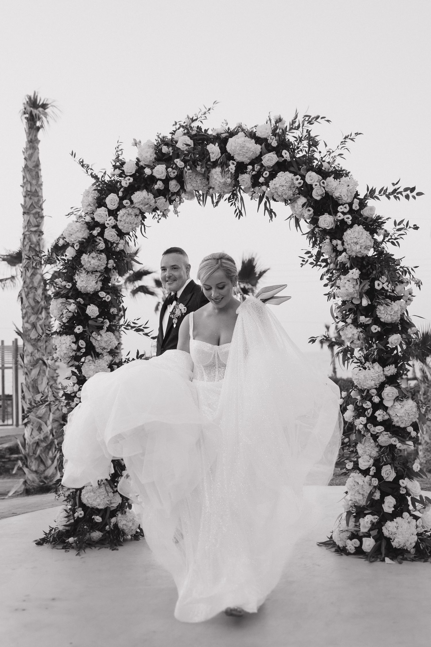 A stunning floral arch frames a wedding couple dancing outdoors, with flowing white dress and palm trees in the background.