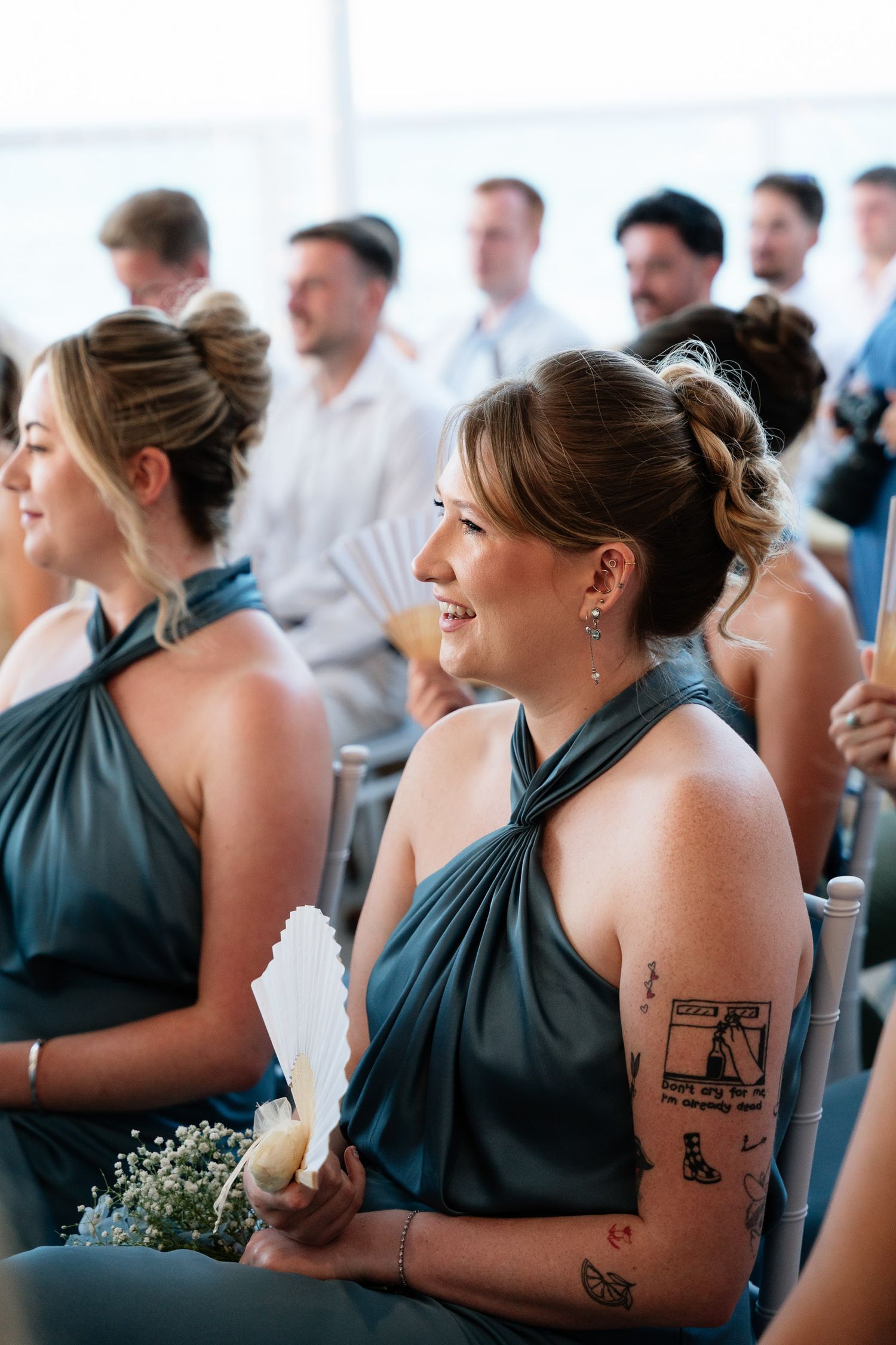 Bridesmaids in charcoal gray dresses laugh together during a wedding celebration, showing matching arm tattoos.