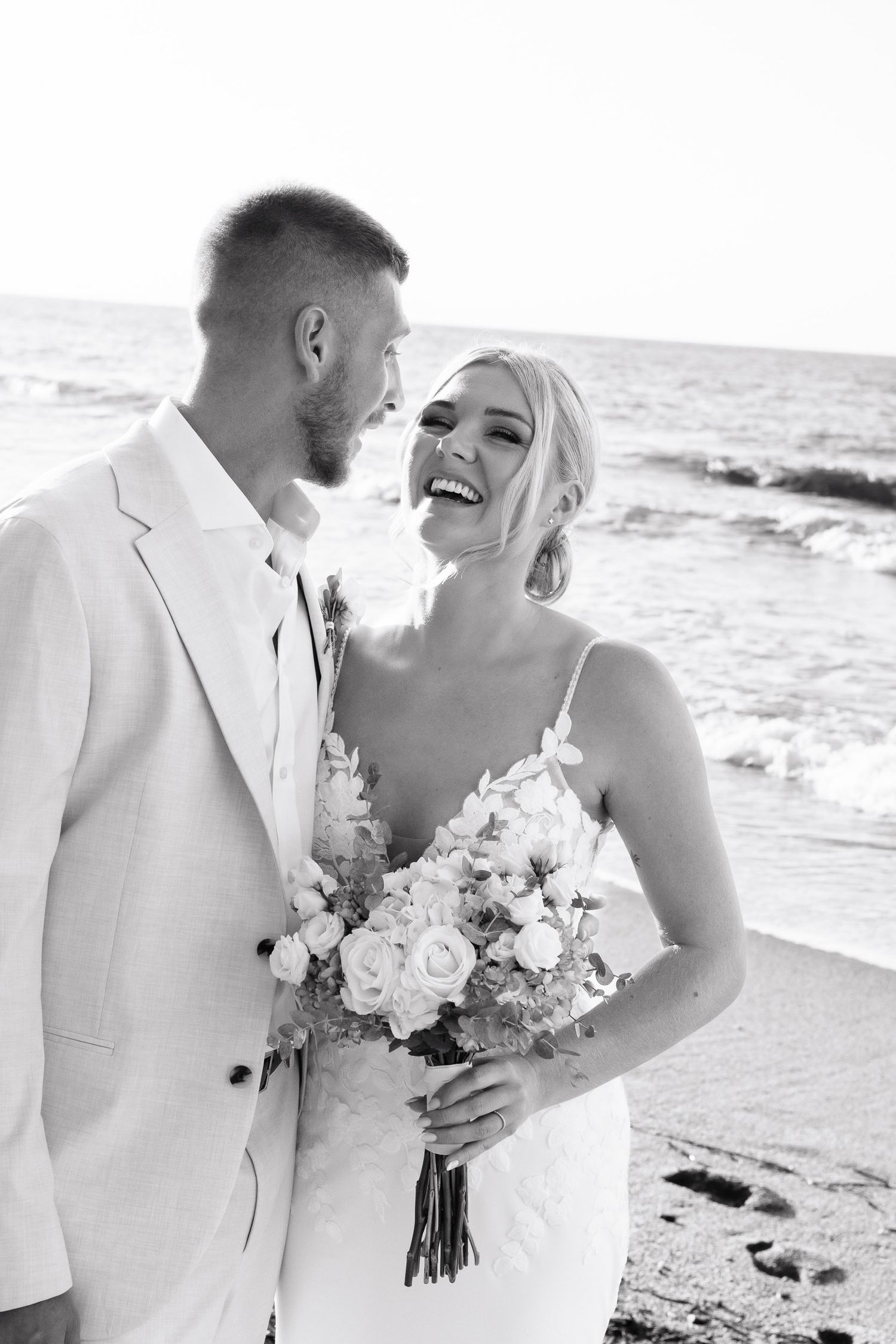 Couple in white attire share intimate moments on a beach with ocean waves in the background.