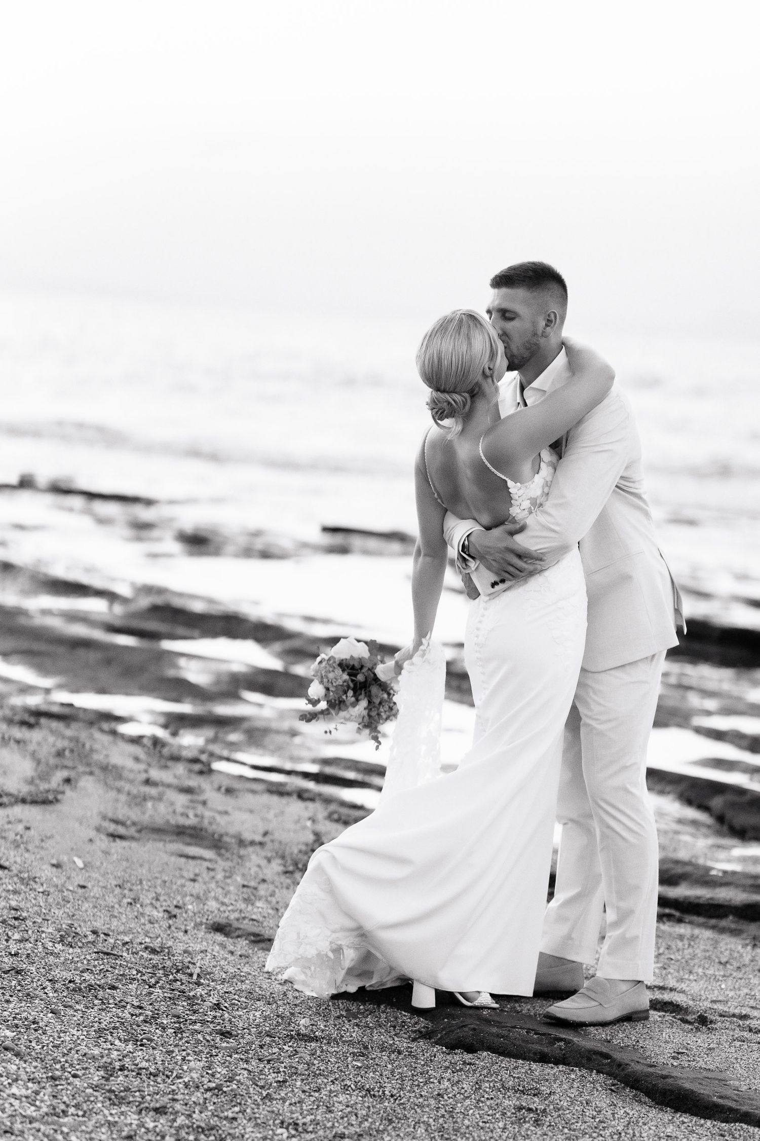A romantic black and white photo series of a couple in wedding attire embracing on a rocky beach.